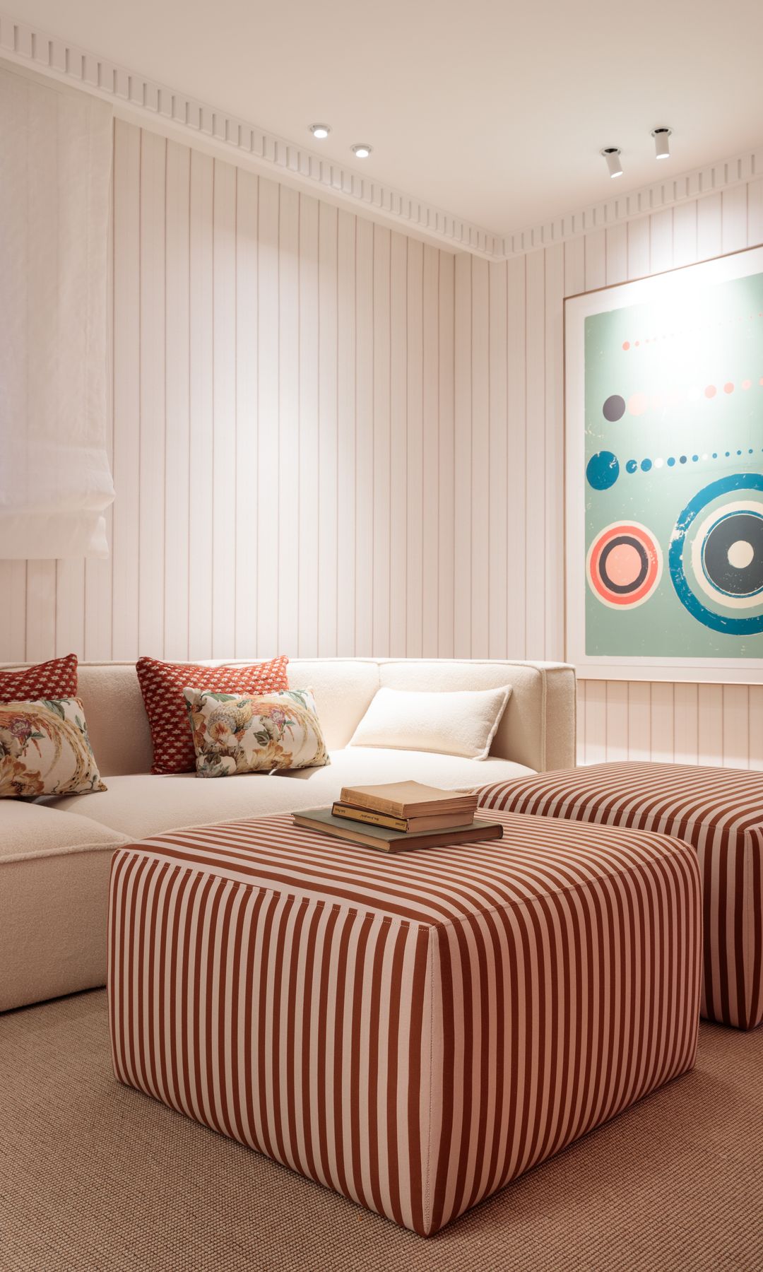 Salón con sofá blanco, póster en la pared de colores, lamas de madera en la pared, pufs de rayas blancas y rojas que son también mesas de centro tapizadas 
