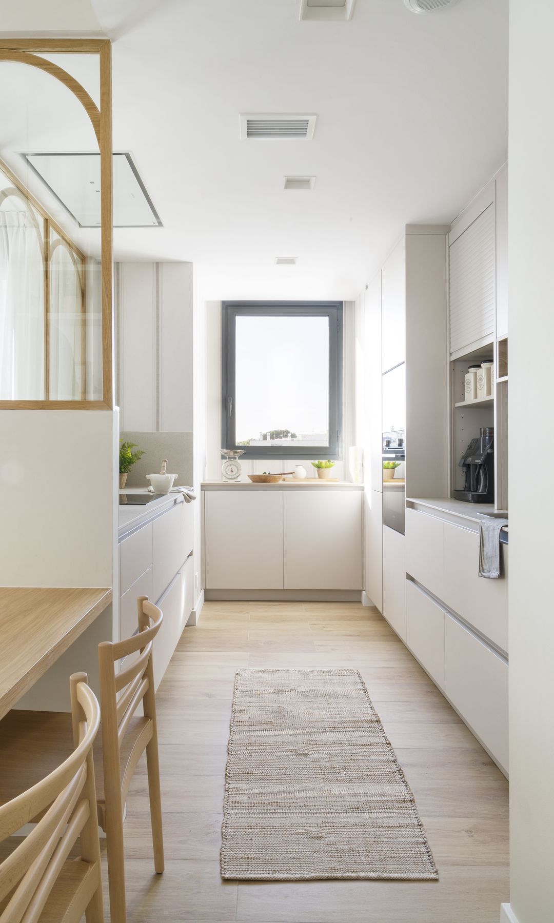 Cocina alargada blanca, suelo de madera, alfombra tipo pasillero, muebles hasta el techo, ventana con el marco negro