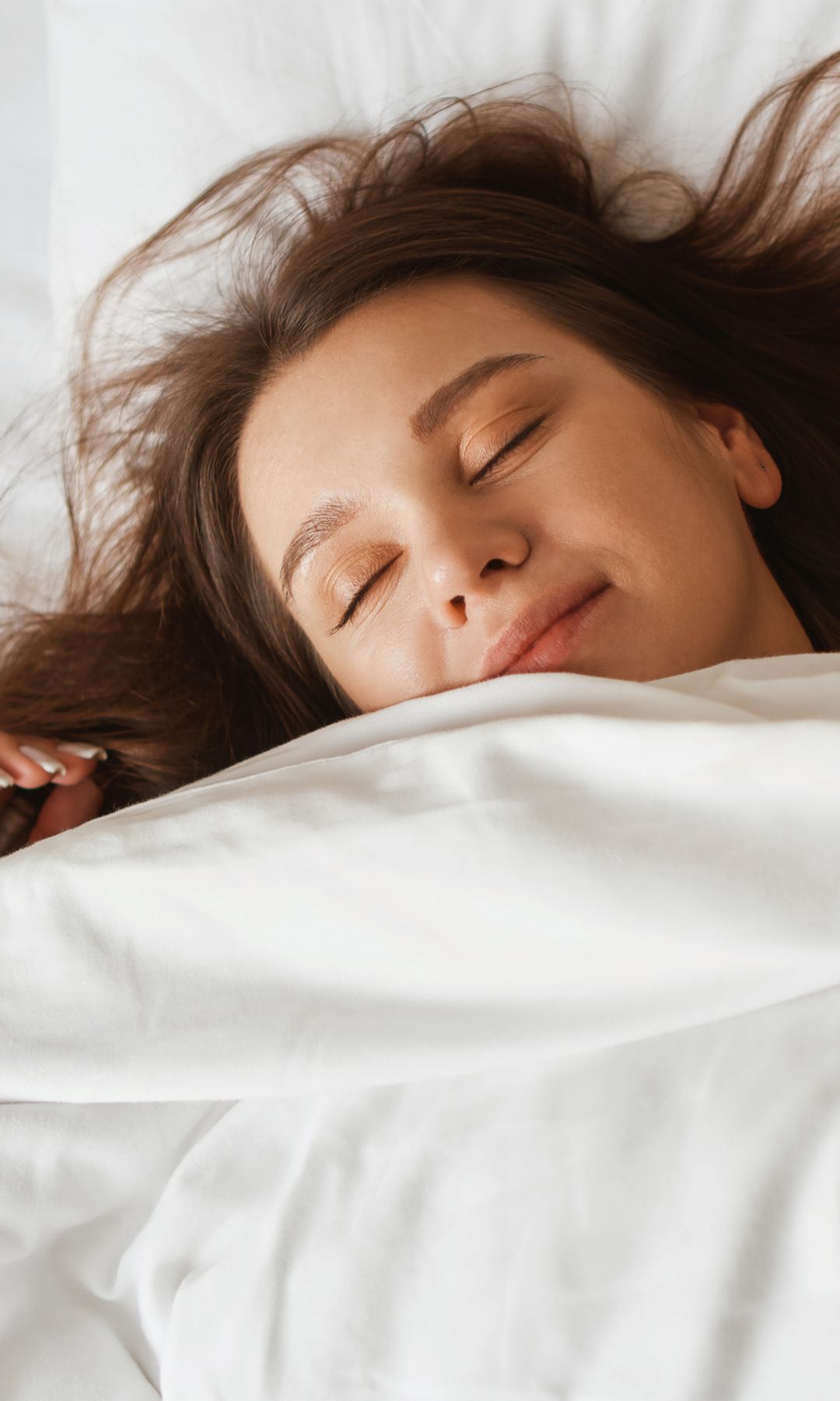 Mujer joven durmiendo en una cama de sábanas blancas con el pelo suelto, boca arriba y sonriente.