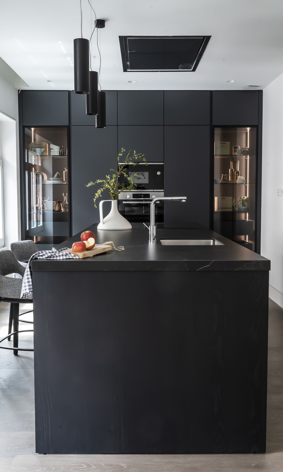 Cocina con isla y muebles negros, lámparas de techo, campana de techo y sillas tapizadas
