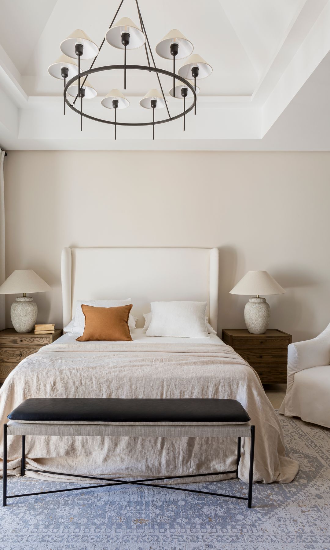 Dormitorio con cabecero tapizado en blanco, techo con molduras, lámpara de techo, alfombra, mesitas de noche de madera, lámparas de sobremesa blancas, banco a los pies de la cama