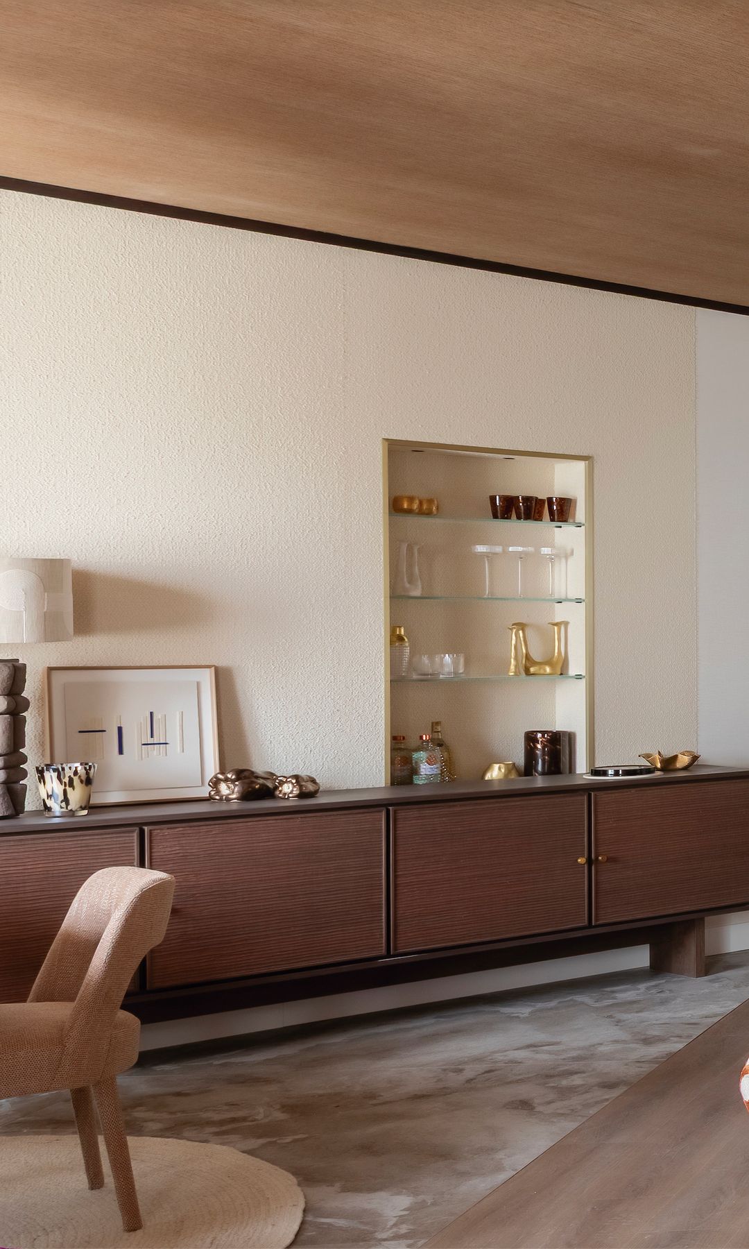 Salón con sofá naranja, aparador bajo de madera oscura, papel pintado que imita el gotelé, hornacina con mueble bar y baldas de cristal, espejo redondo negro 