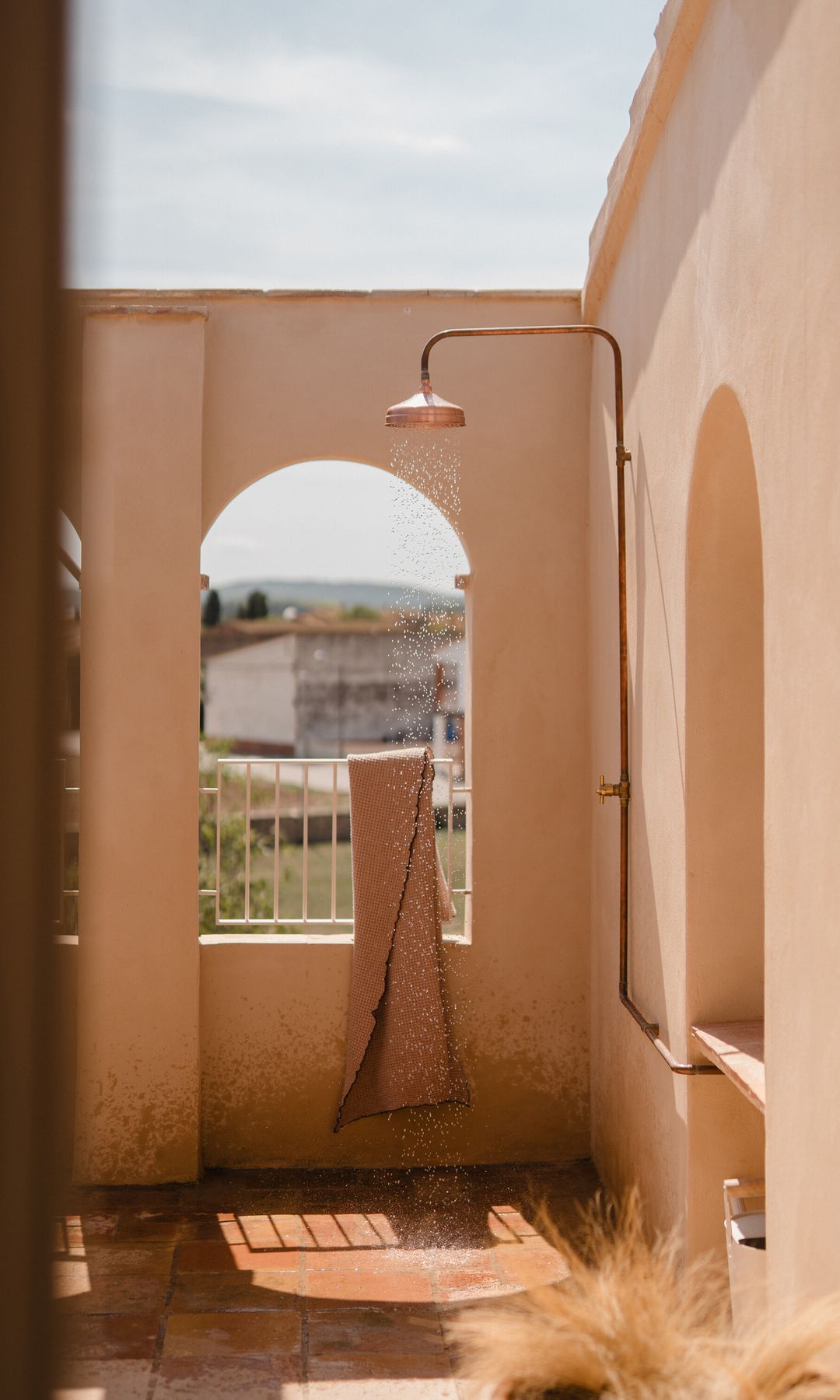 Ducha en terraza con suelo de barro y alcachofa de color cobre, toalla en la barandilla 