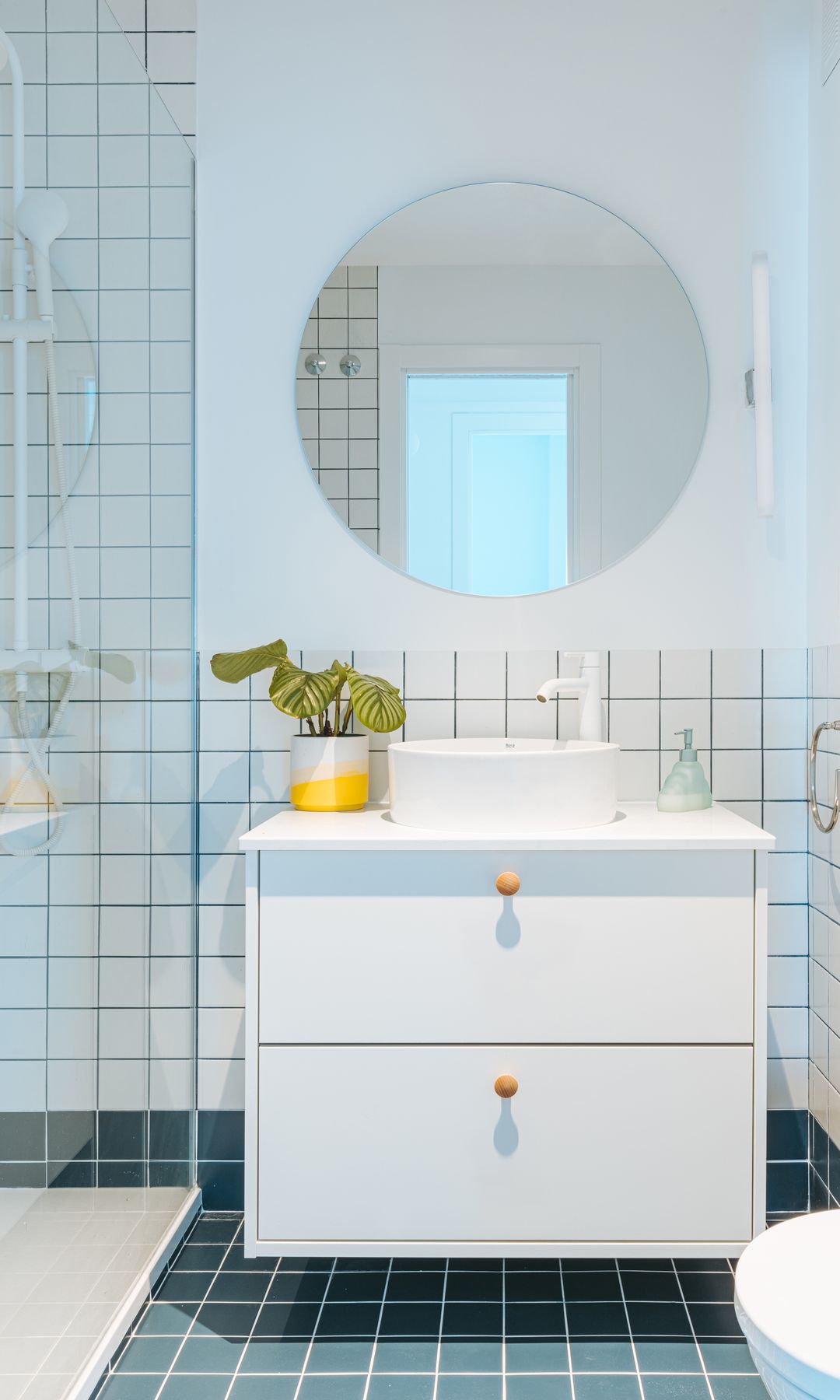 Baño con zócalo alto de azulejos blancos cuadrados, suelo de azulejos azules, espejo redondo, ducha con mampara, mueble de lavabo suspendido