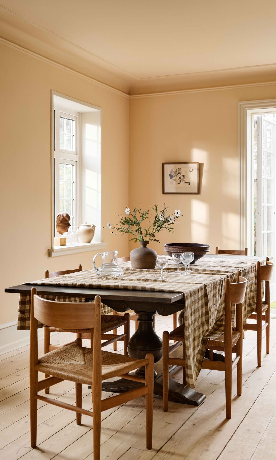 Comedor con paredes en color crema cálido, mesa de comedor de madera oscura, sillas de madera con el asiento de enea, mantel de cuadros