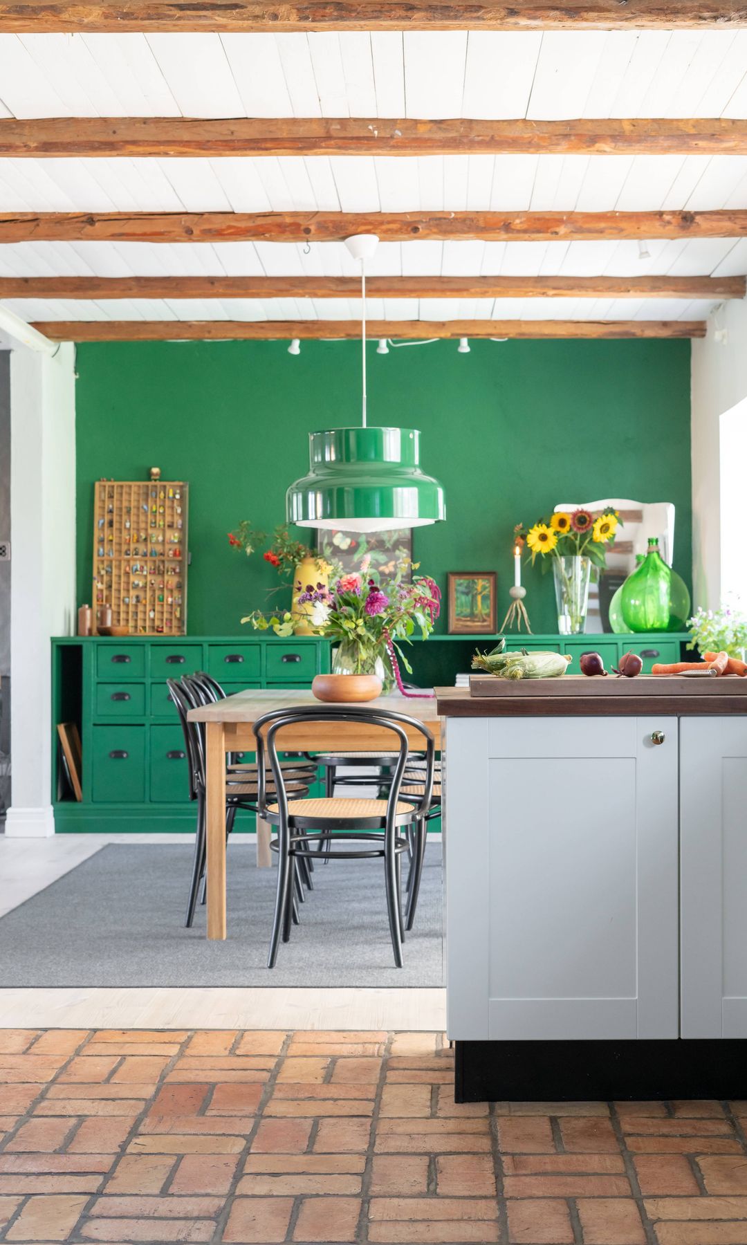 Cocina con mesa de cocina, suelo de barro, vigas de madera, pared en verde botella, mueble bajo a juego, lámpara de techo verde sobre la mesa de cocina 