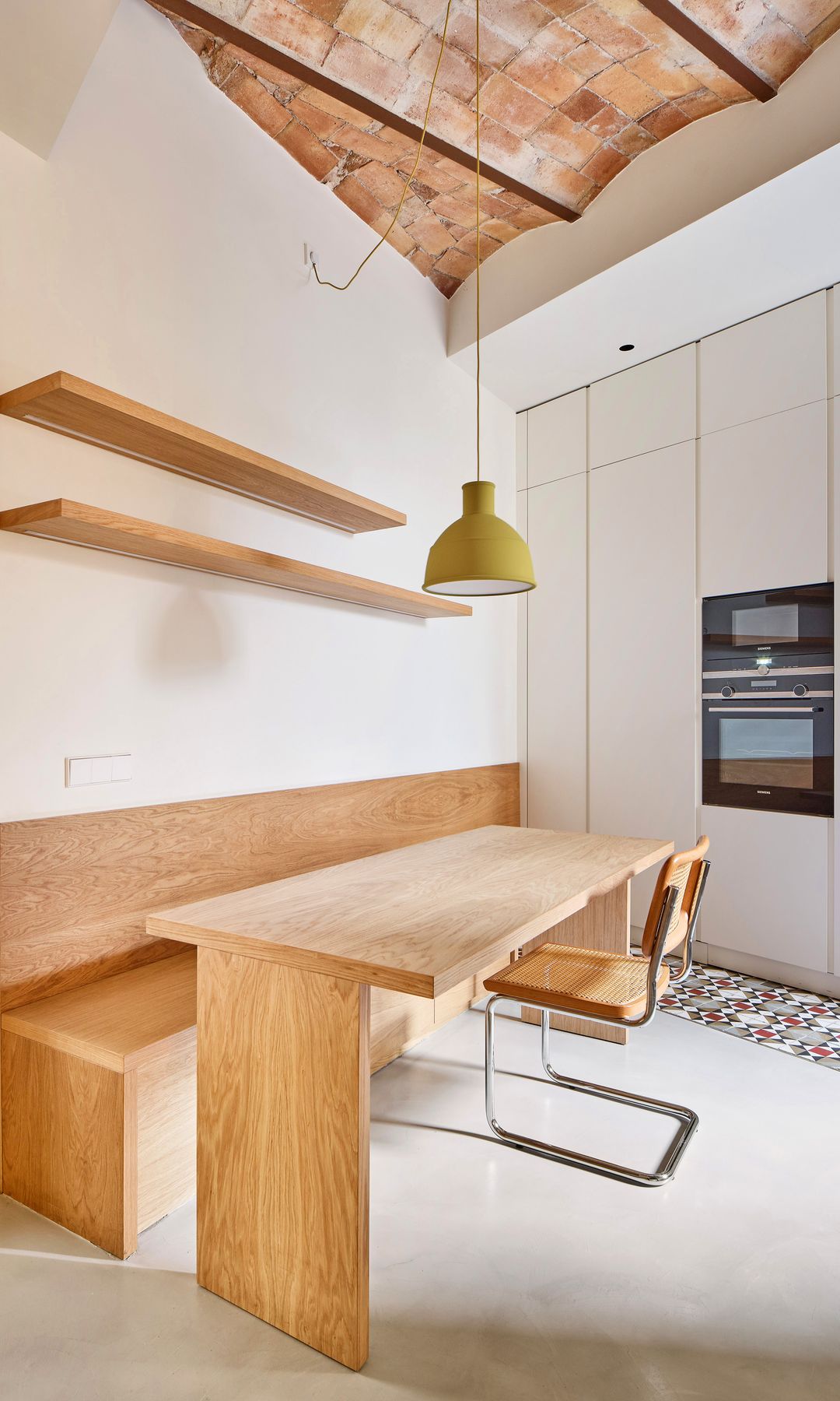 Cocina con comedor, mesa de madera, banco de madera, baldas, muebles blancos, bóveda catalana, horno y microondas en columna