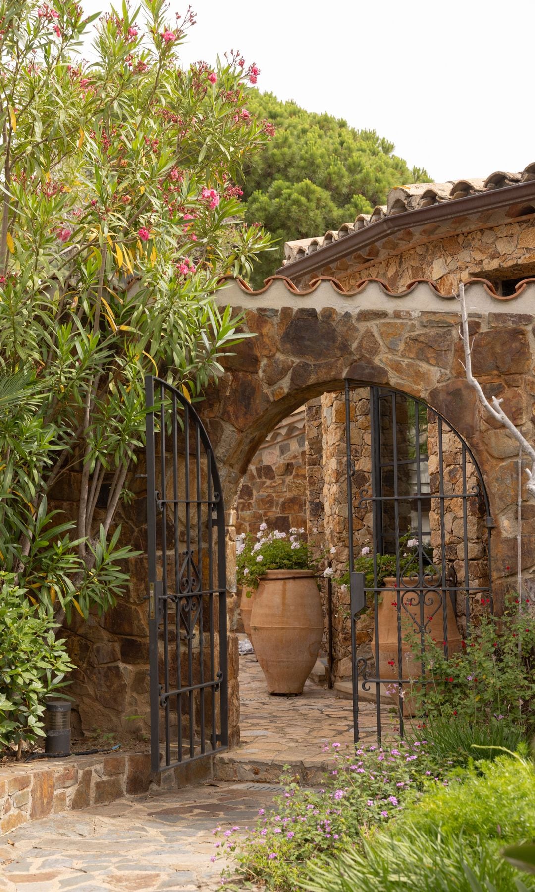 Entrada casa de piedra con grandes macetas con flores, puerta de hierro en forma de arco