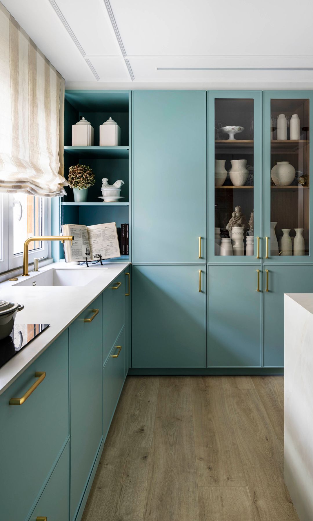 Cocina con vitrina y muebles en color azul claro, estores en las ventanas, suelo de madera, tiradores y grifería doradas 