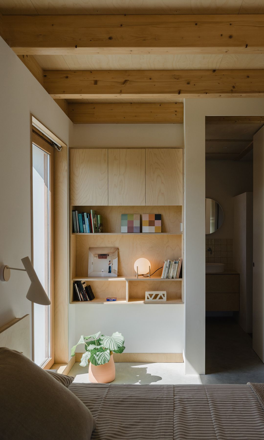 Rincón dormitorio con estantería a medida de madera, ventanal, maceta en el suelo, libros y lámpara de sobremesa 