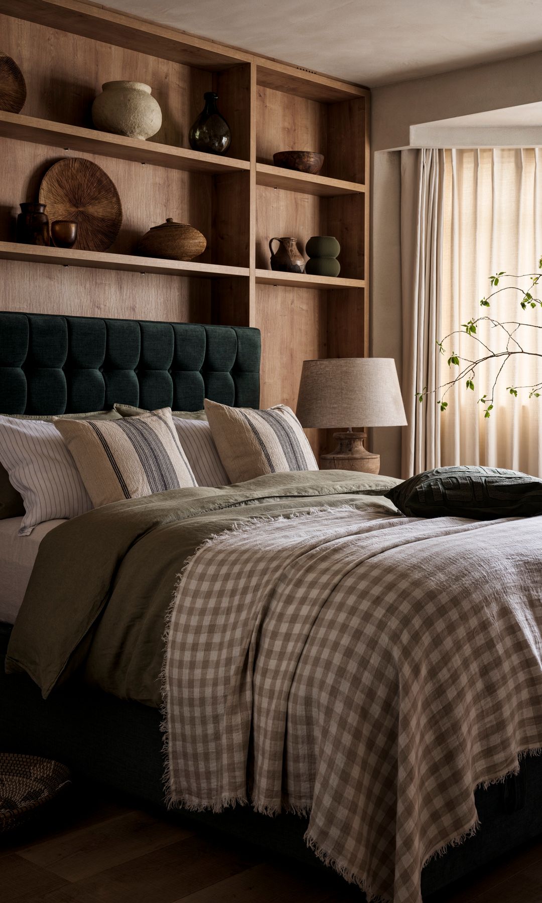 Dormitorio con pared del cabecero con estantería a medida de madera, cabecero de terciopelo, cortinas, ropa de cama de cuadros, lámpara de sobremesa 