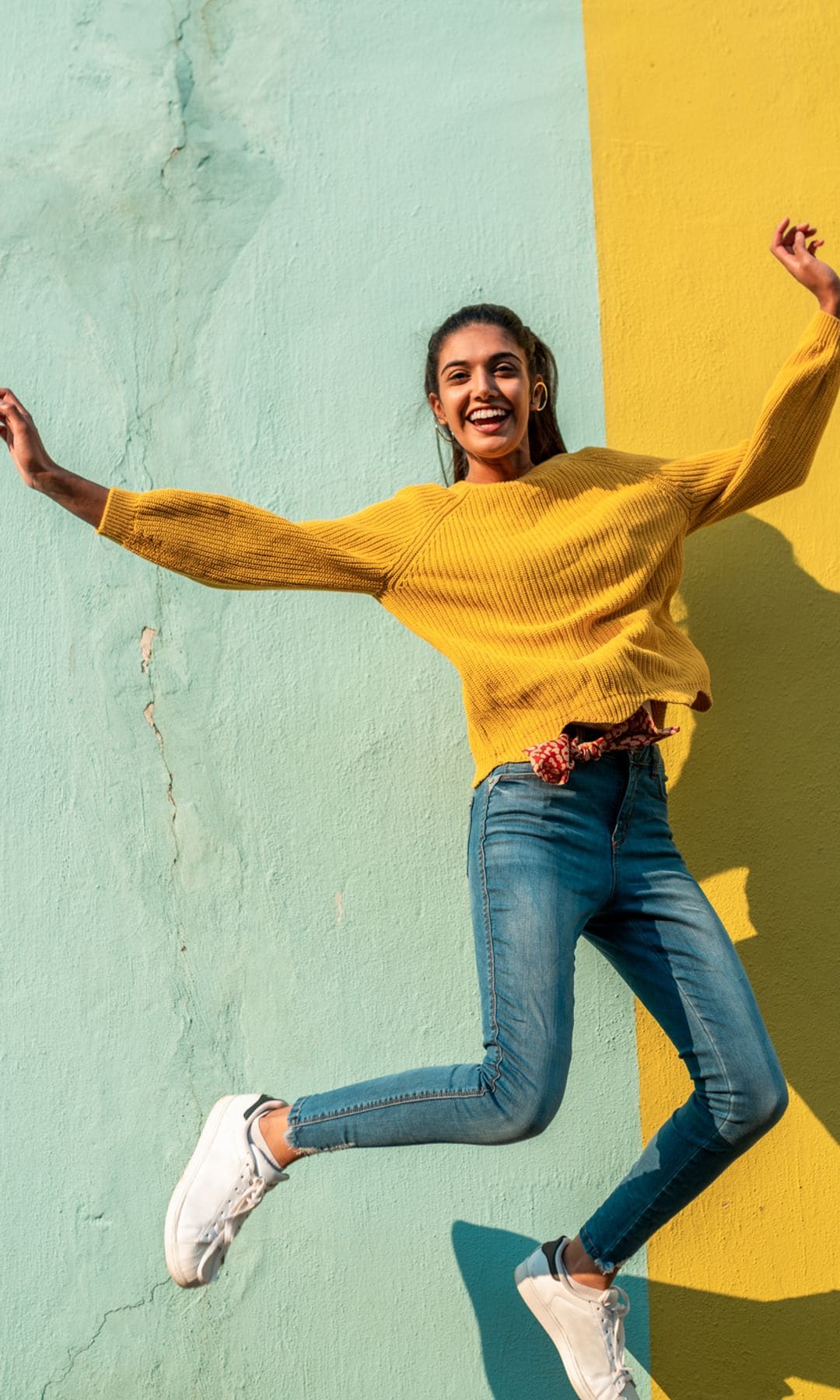 Mujer saltando con alegría frente a una pared amarilla y azul.
