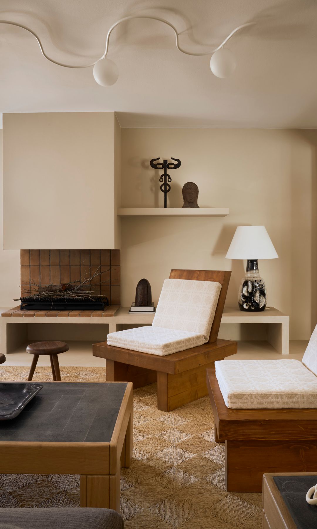 Salón con chimenea y butacas de madera con cojín blanco, mesa de centro con el sobre negro, lámpara de techo, alfombra de fibras vegetales y lámpara de sobremesa 