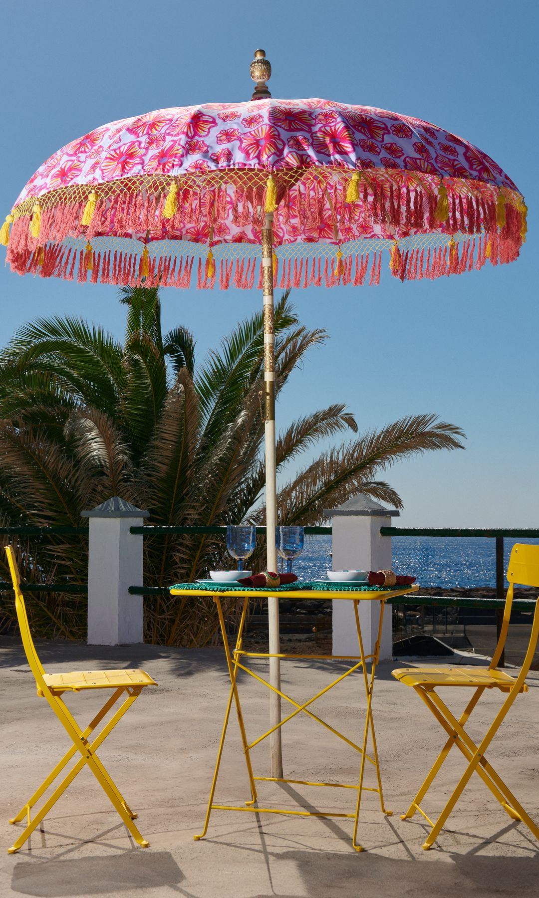 Terraza con silla y mesas de hierro pintado de amarillo, sombrilla con flecos en rosa y amarillo