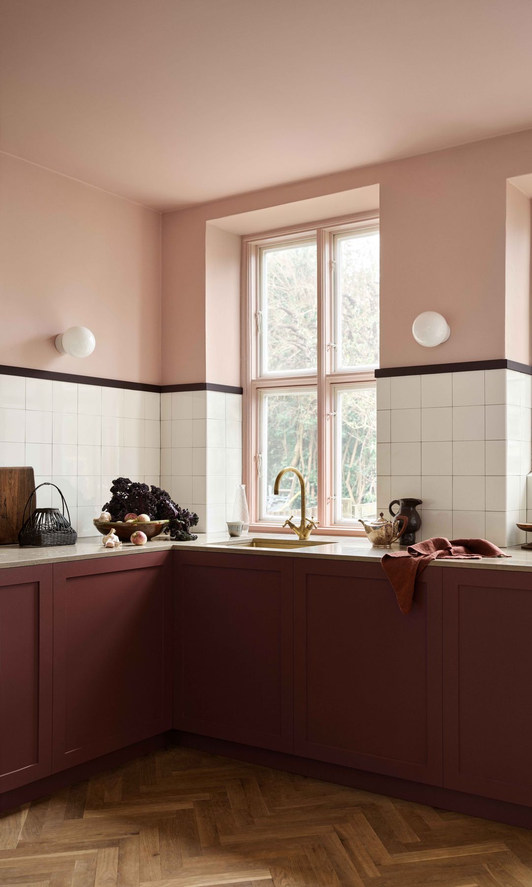 Cocina con el suelo y las paredes en color rosa palo, zócalo alto de azulejos, muebles burdeos, apliques de pared, suelo de madera