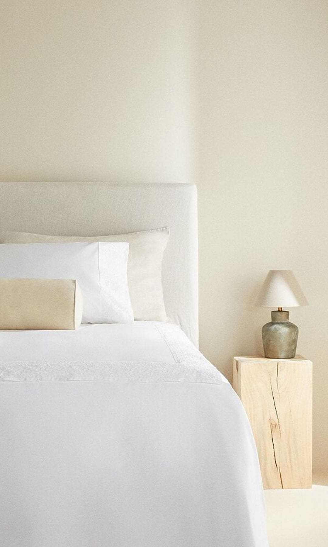 Cama en blanco con una mesilla de madera y una pequeña lámpara sobre ella. Todo muy iluminado y muy blanco.