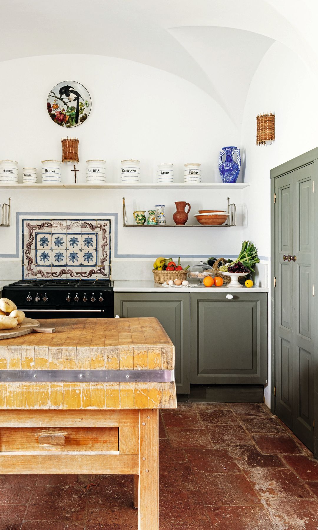Cocina rústica con mesa de madera, estantes con jarras, y frutas en la encimera.