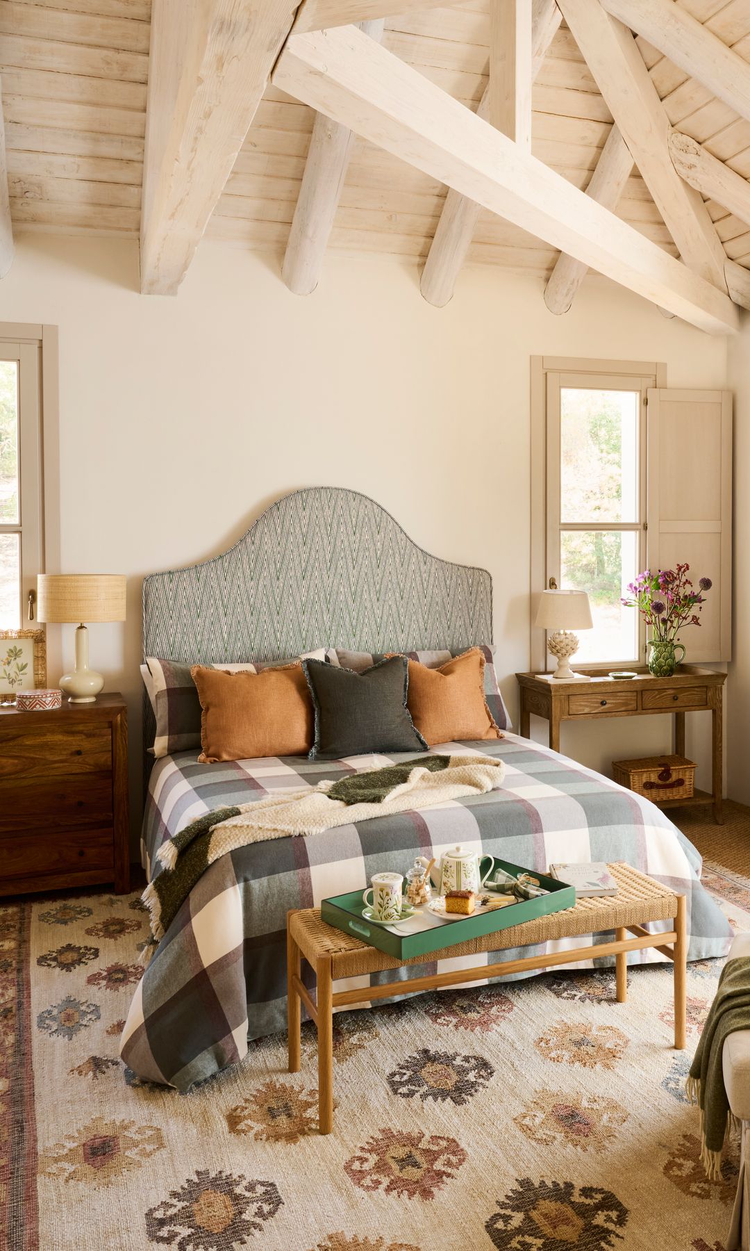 Dormitorio abuhardillado con vigas de madera blanca, alfombra, cabecero tapizado en ondas, ropa de cama de cuadros, cómoda haciendo de mesita de noche, consola 