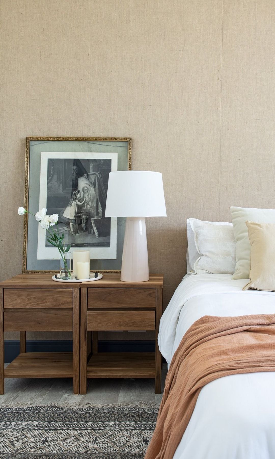 Dormitorio con mesita de noche doble de madera, lámpara de sobremesa blanca, fotografía enmarcada apoyada en la mesita de noche, vela y jarrón pequeño con flores 