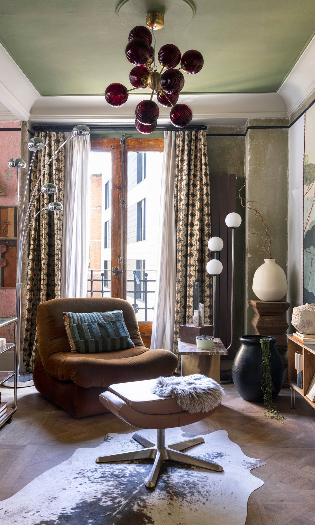 Salón con papel pintado en la pared, techo verde con moldura, sillón marrón, reposapiés de cuero, alfombra de piel de vaca, cortinas y visillos, estantería baja, lámpara de techo