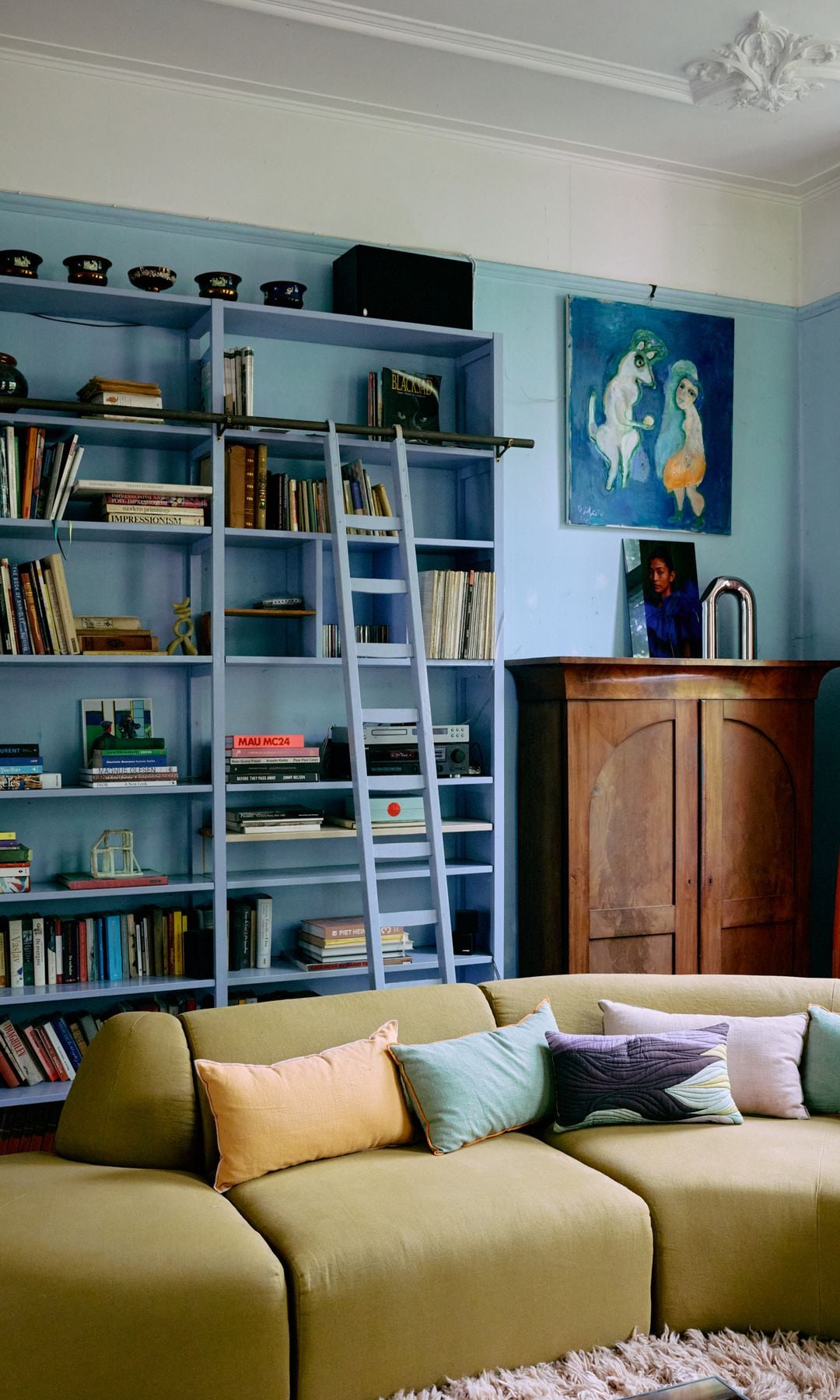 Salón con paredes y estantería de color azul, sofá en color mostaza, alfombra de pelo largo, escalera, molduras en el techo, aparador de madera con una foto encima