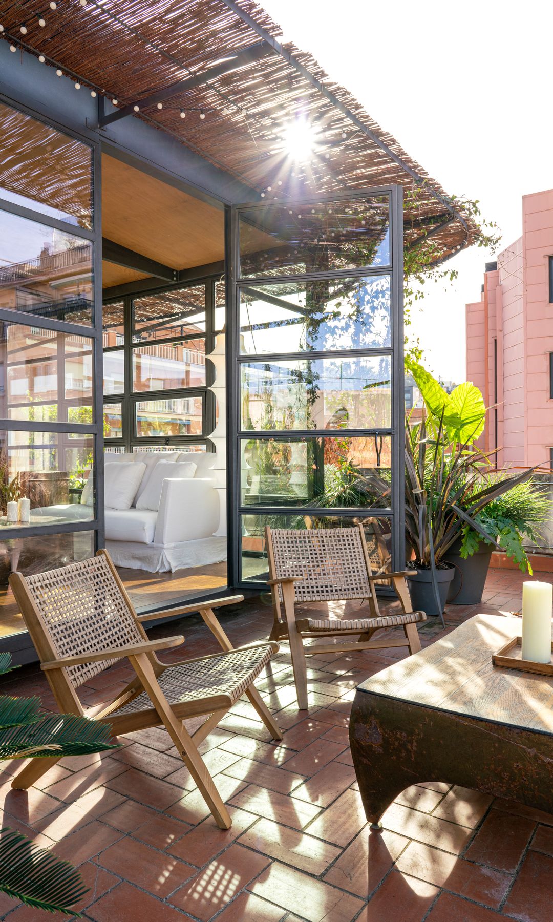 Terraza con entrada a la casa con grandes puertas de cristal, mesa de madera, suelo de barro y butacas de fibras