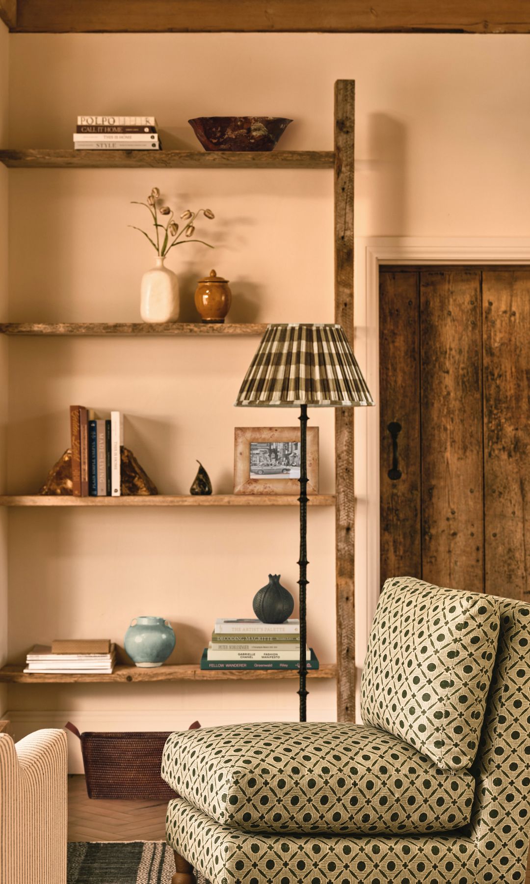 Salón con baldas de madera, fotos y libros,  paredes en color arena, alfombra, butaca, lámpara de pie con pantalla de tela, suelo de madera en espiga