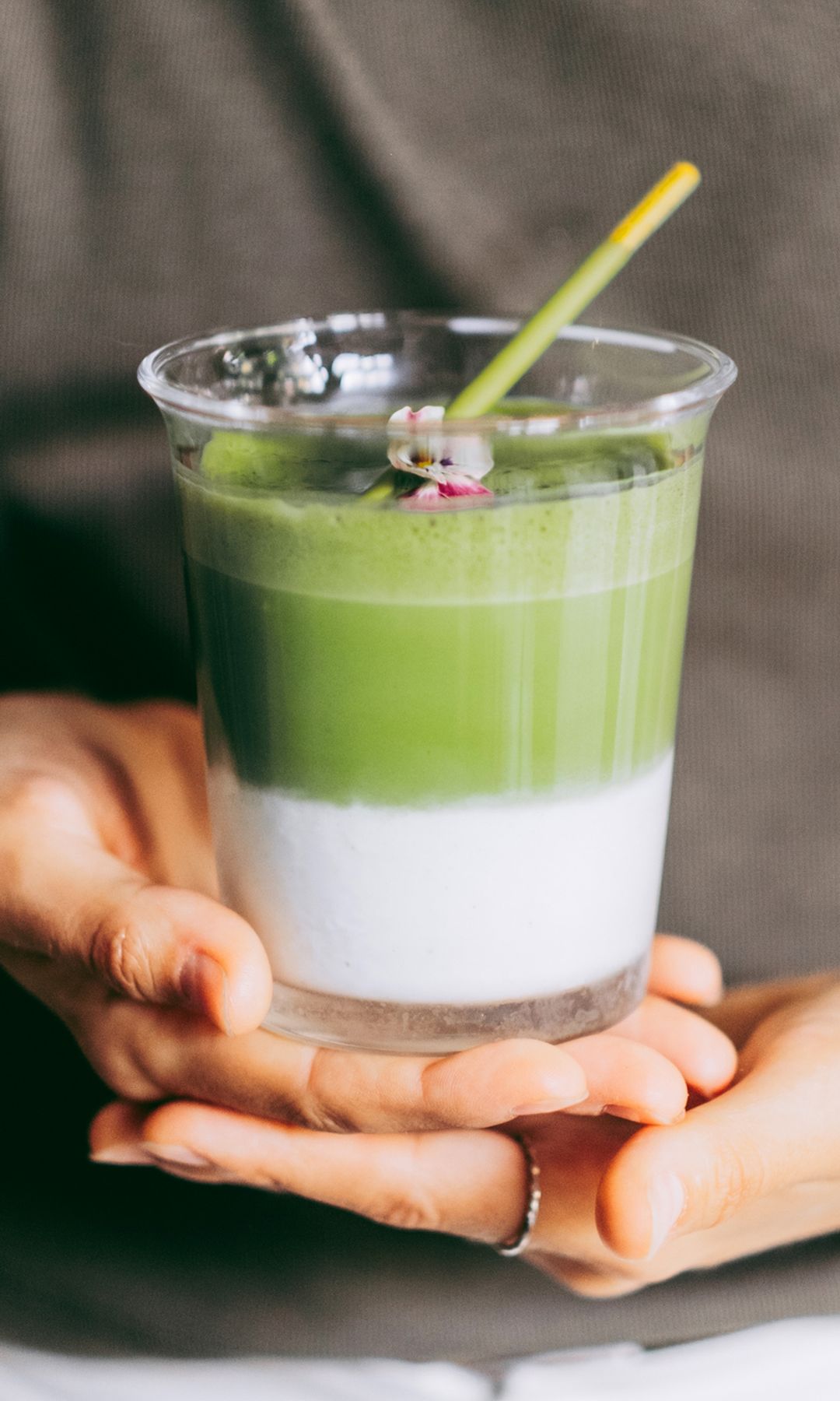 Un vaso de té matcha con leche en las manos de una chica que lleva una camiseta marrón y pantalones blancos. Es un plano detalle del vaso con una pajita.