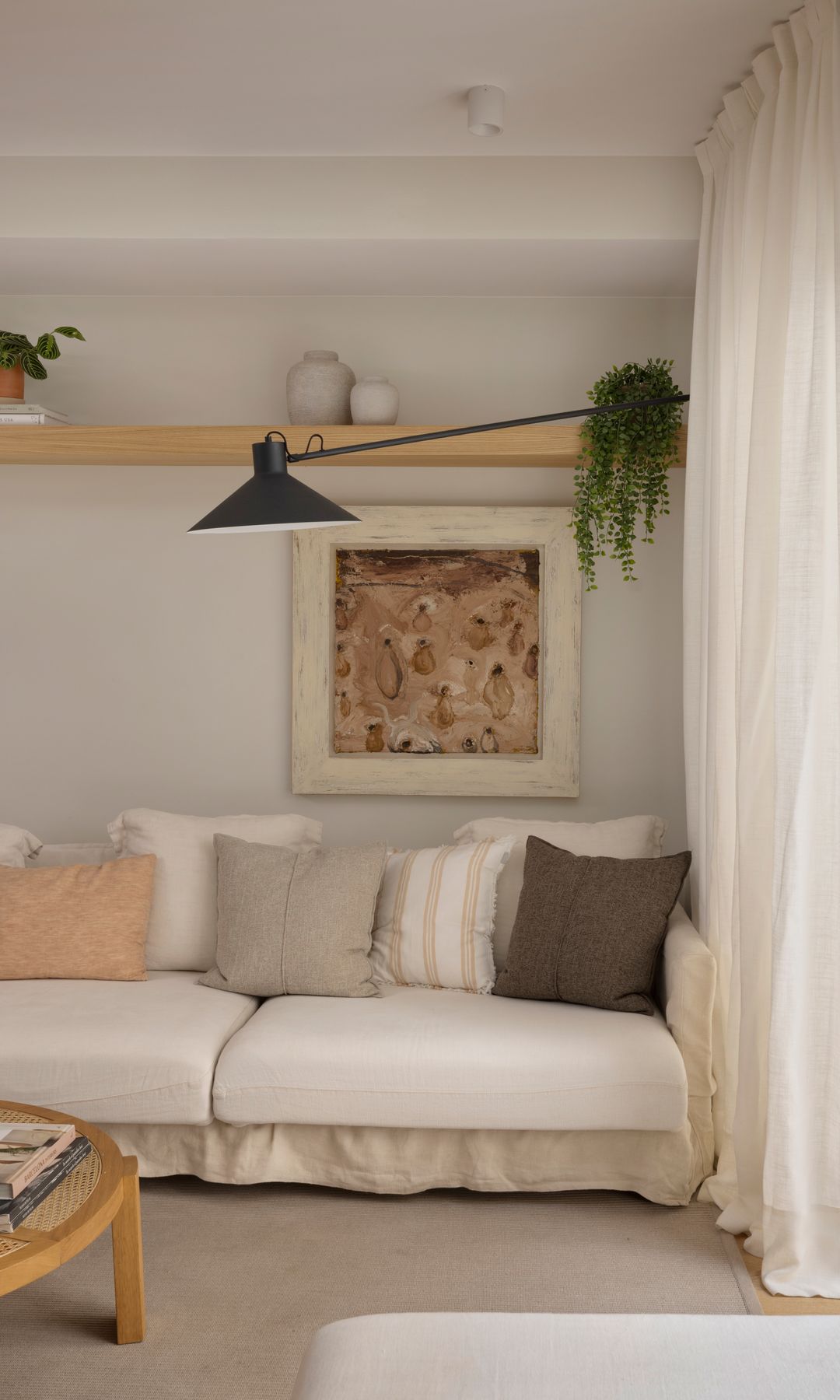 Sofá blanco con volante inferior, cuadro, cojines de diferentes colores, cortinas de lino blancas, alfombra beige, balda de madera sobre el sofá con plantas y adornos  