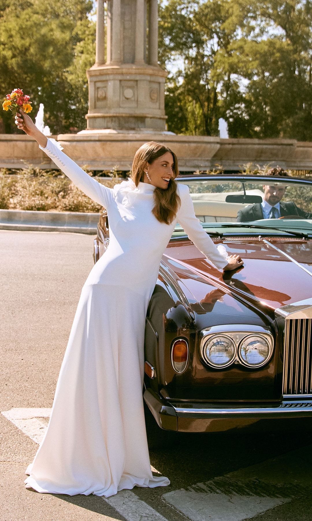 Amor sobre ruedas: Entallado, de manga larga y con cuello alto. Así es este vestido con semivuelo de María Barragán, que en esta imagen se acompaña de pendientes y sortija de oro blanco y diamantes, de Rabat. Él, con total look, de Budiman, conduce un exclusivo -y único- Rolls Royce Corniche burdeos del año 1985, con tapicería en color magnolia. Es de la firma Coches Únicos by Nacho de Villota