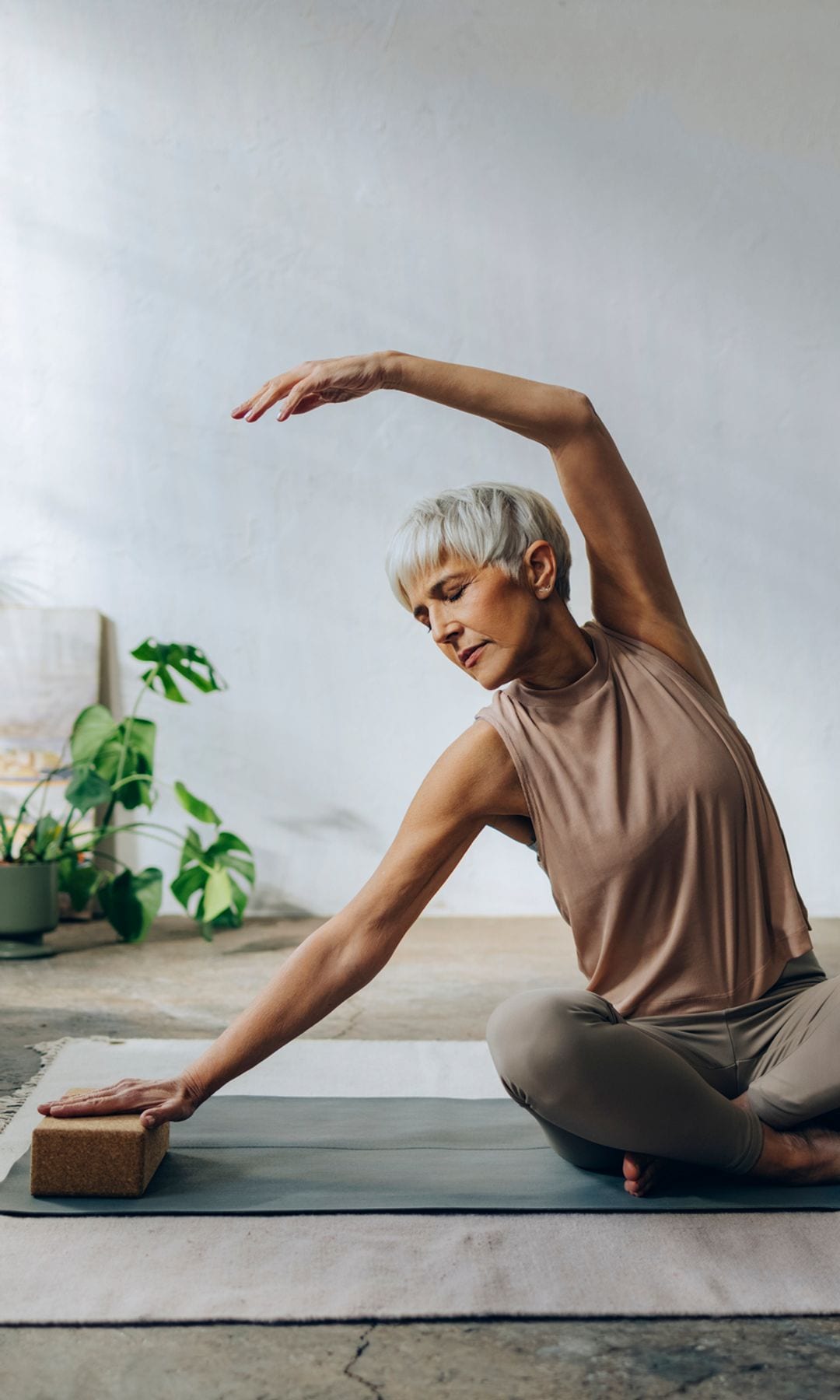 Mujer a partir de 50 años practicando yoga sobre una esterilla, con un bloque, en el suelo de su casa.