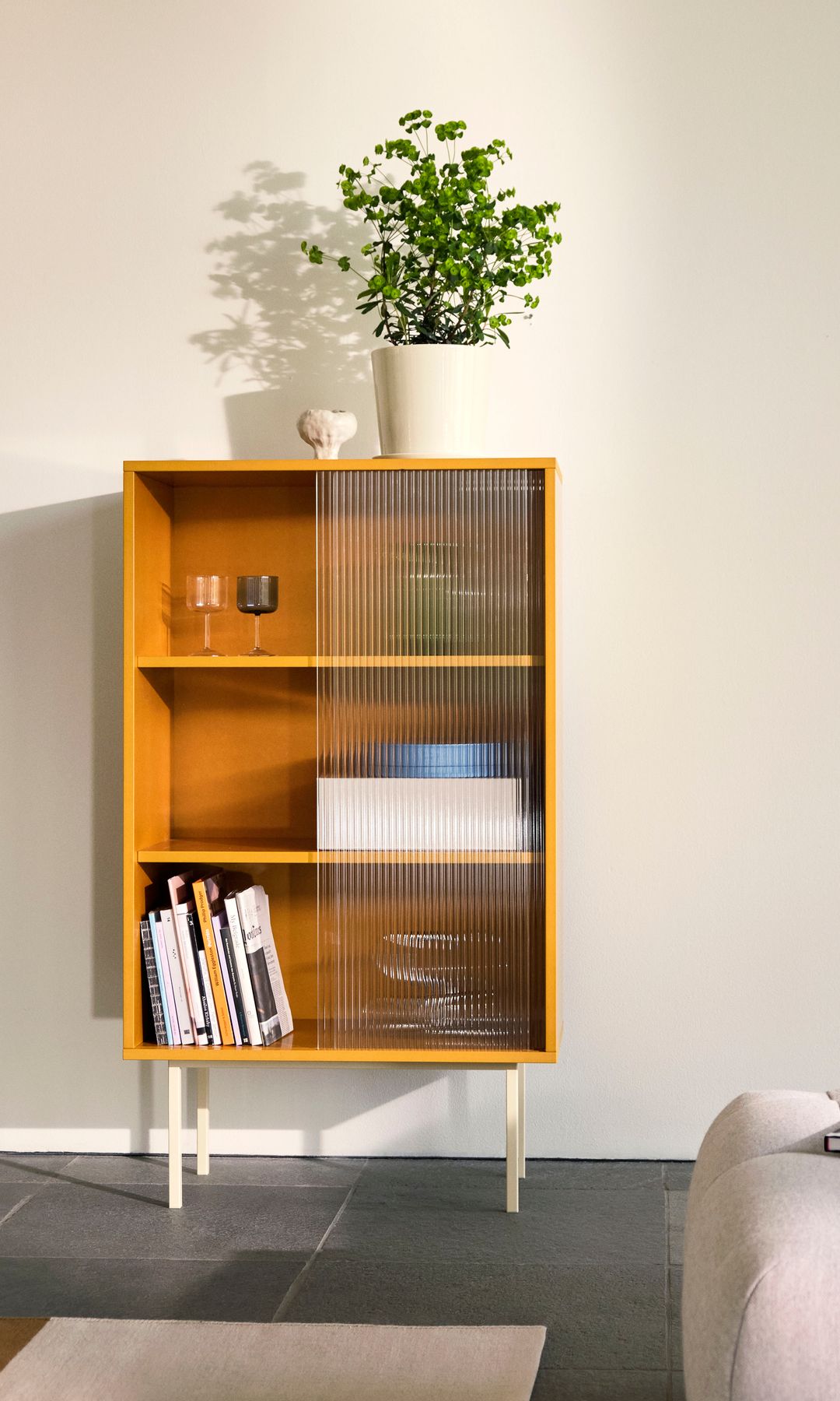 Vitrina de color amarillo y puertas de cristal esmerilado con patas blancas, maceta con planta en la parte superior