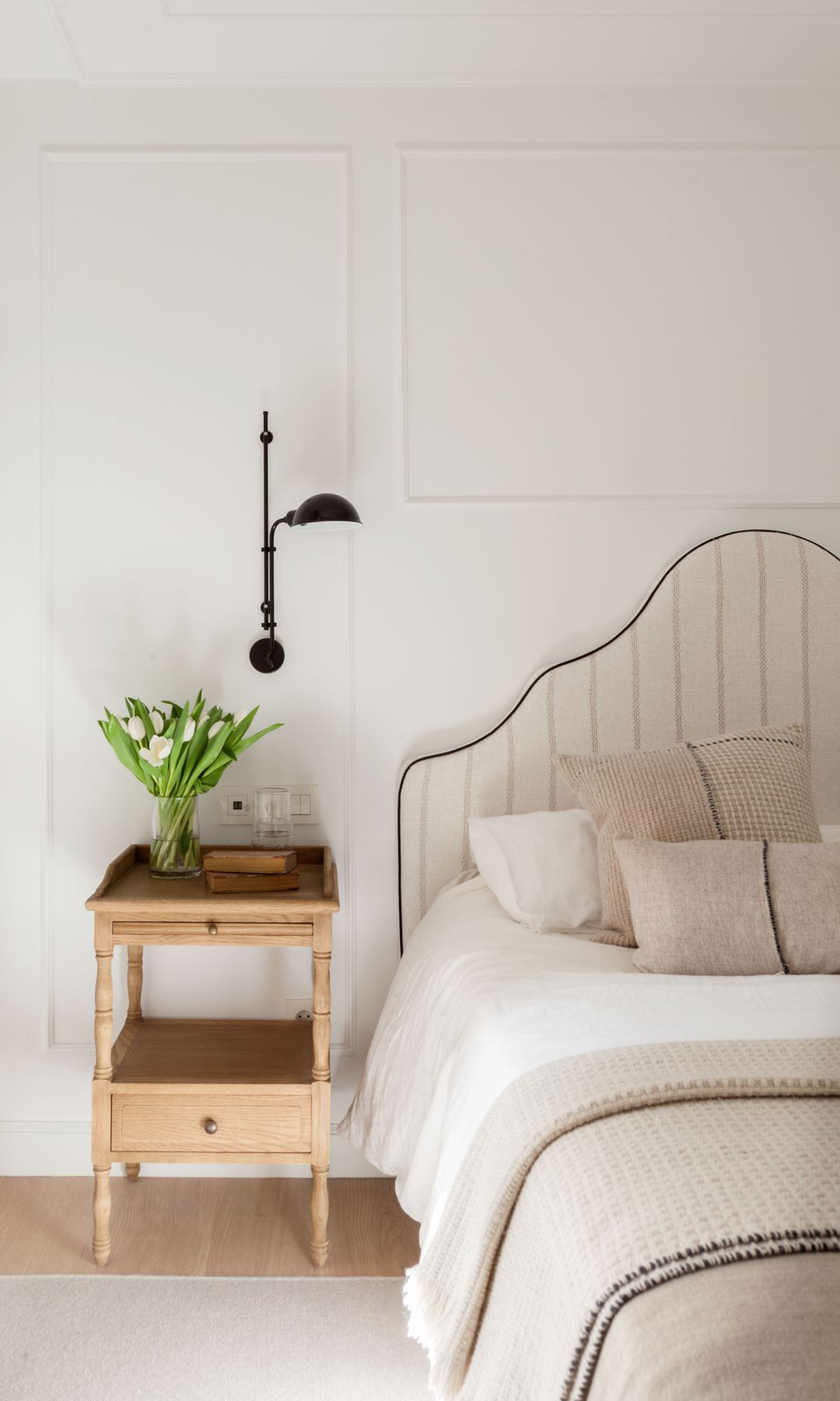 Dormitorio con cabecero tapizado tipo pagado, mesita de noche de madera, aplique de pared, jarrón con tulipanes 