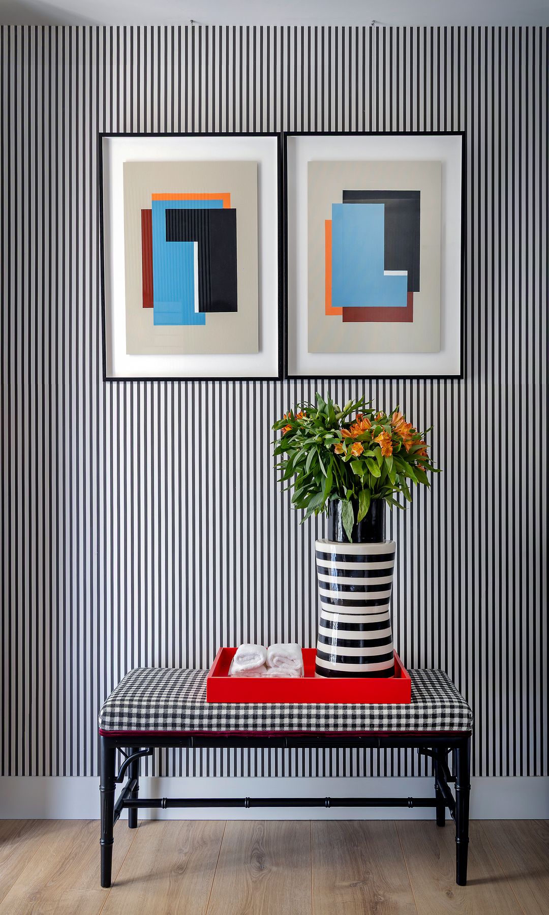 Rincón con papel pintado a rayas verticales finas negras y blancas, butaca tapizada con cuadros blancos y negros, jarrón blanco y negro, cuadros en la pared, bandeja roja 