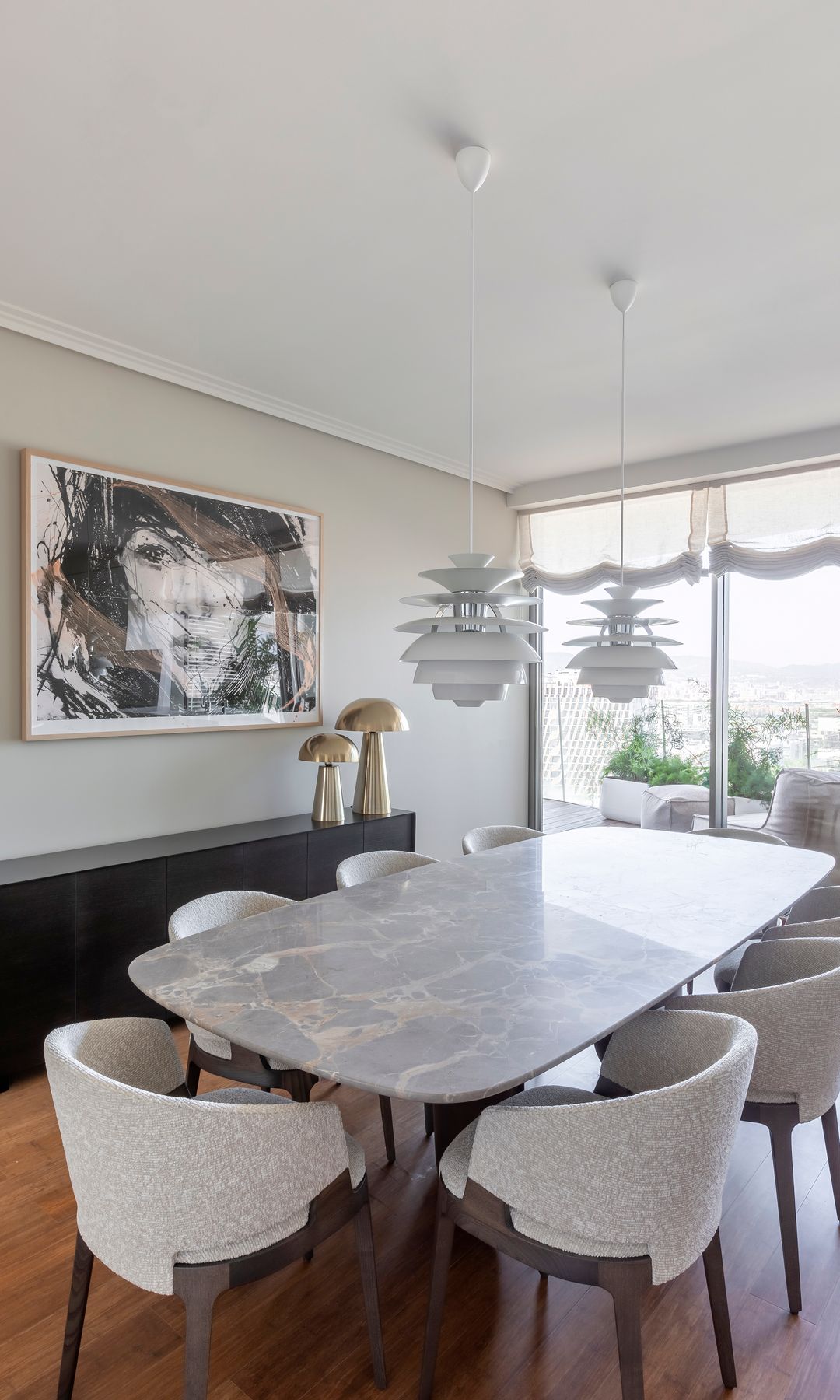 Comedor con mesa ovalada con el sobre de mármol, cuadro en la pared, lámparas de techo blancas, suelo de madera, aparador negro, estores blancos 
