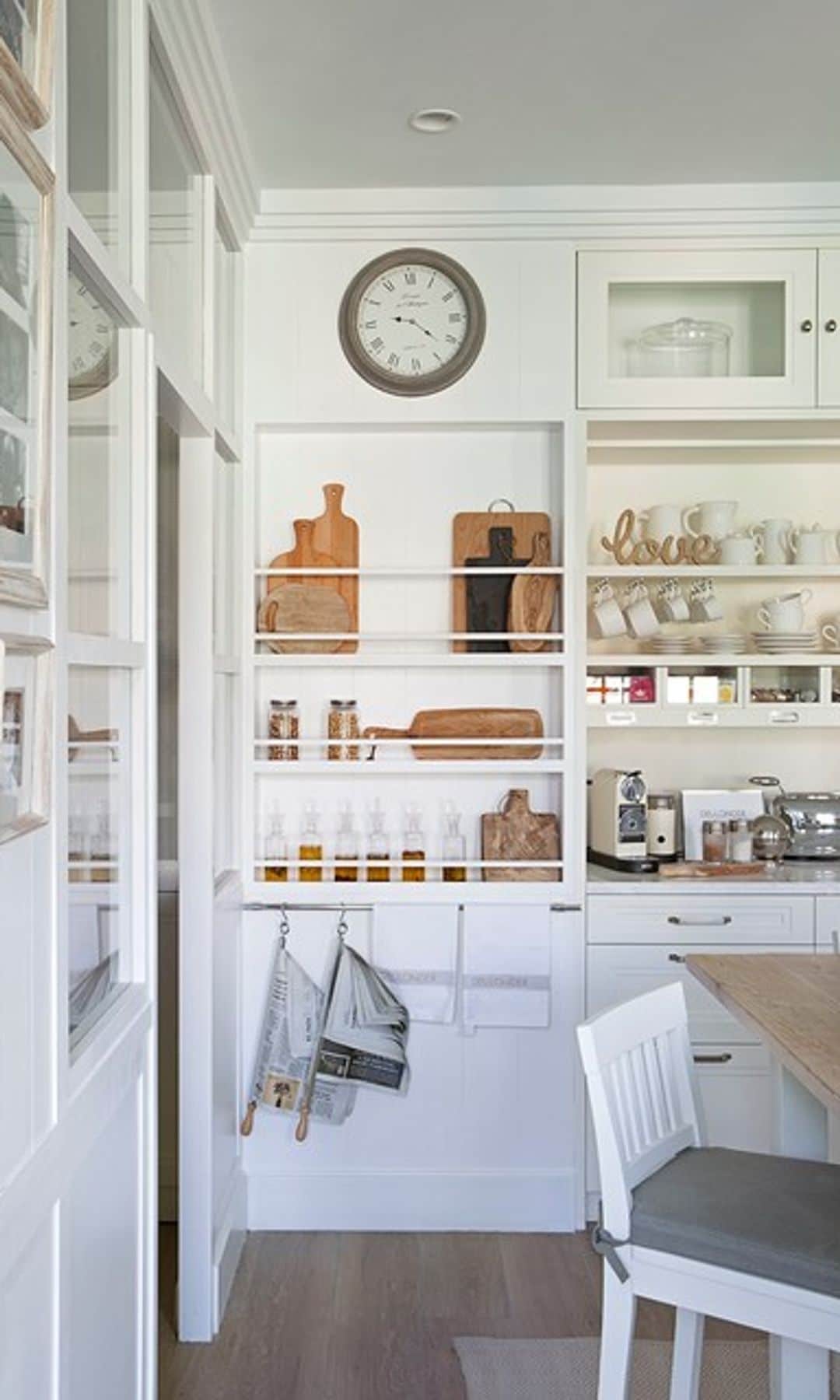 Cocina blanca con baldas y barras para accesorios, reloj en la pared, paños de cocina en ganchos, especieros, tablas de cortar