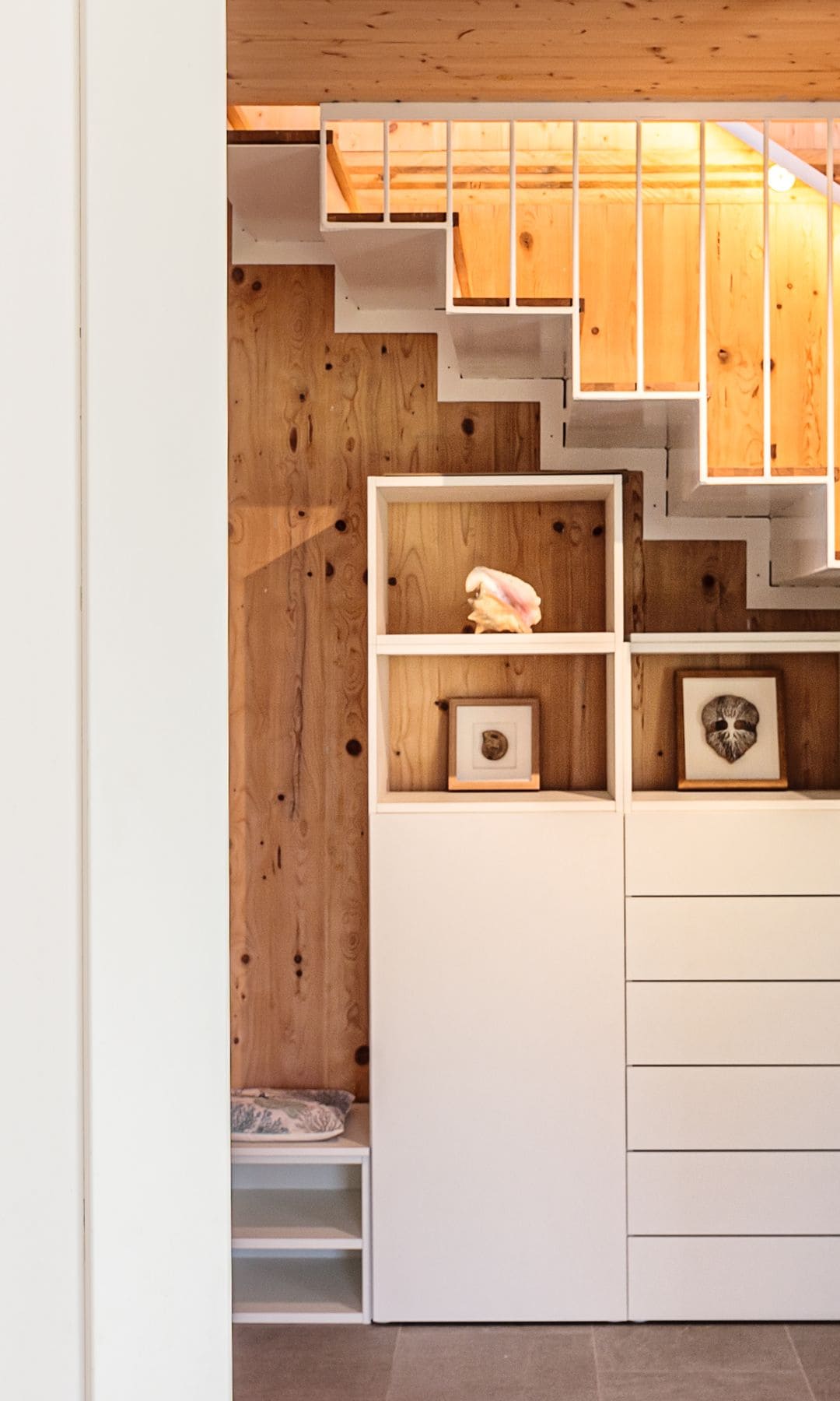 Mueble blanco con huecos abiertos y cajones bajo la escalera, con banco incorporado, pared de madera, cuadros en las baldas, barandilla metálica blanca