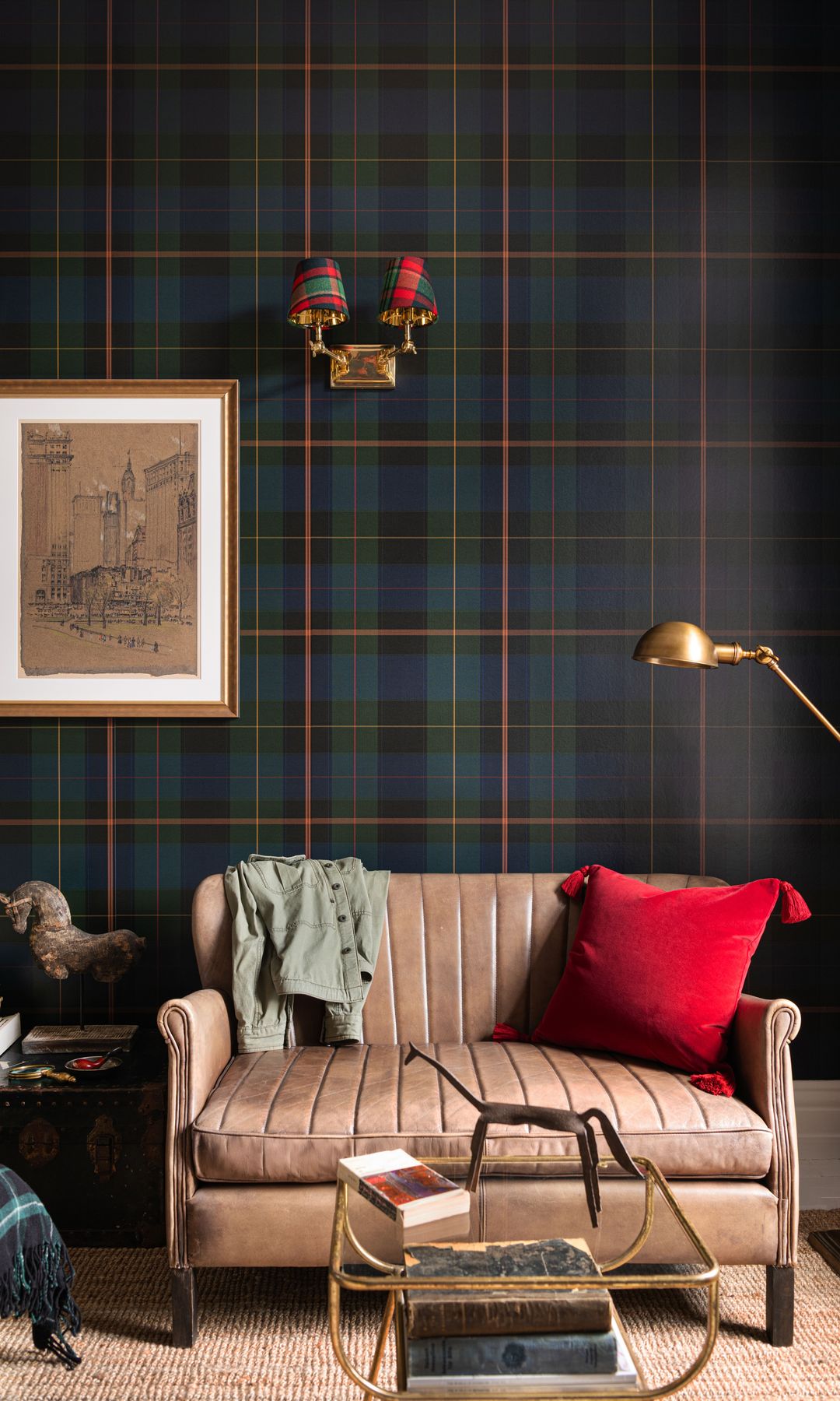 Salón con papel pintado de cuadros tipo tartán, sofá de cuero marrón claro, cojín rojo, lámpara de pie dorada, aplique de pared, cuadro