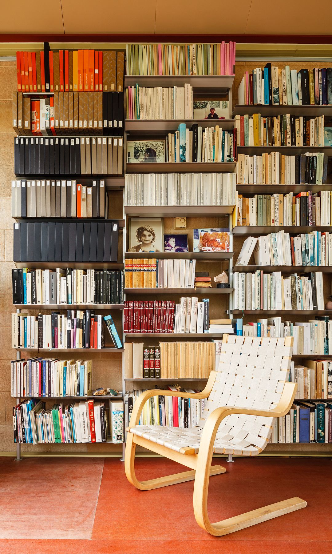 Rincón de lectura con estantería Hypostila de Bd Barcelona llena de libros, pared de azulejos marrones, suelo de baldosas, silla con brazos 