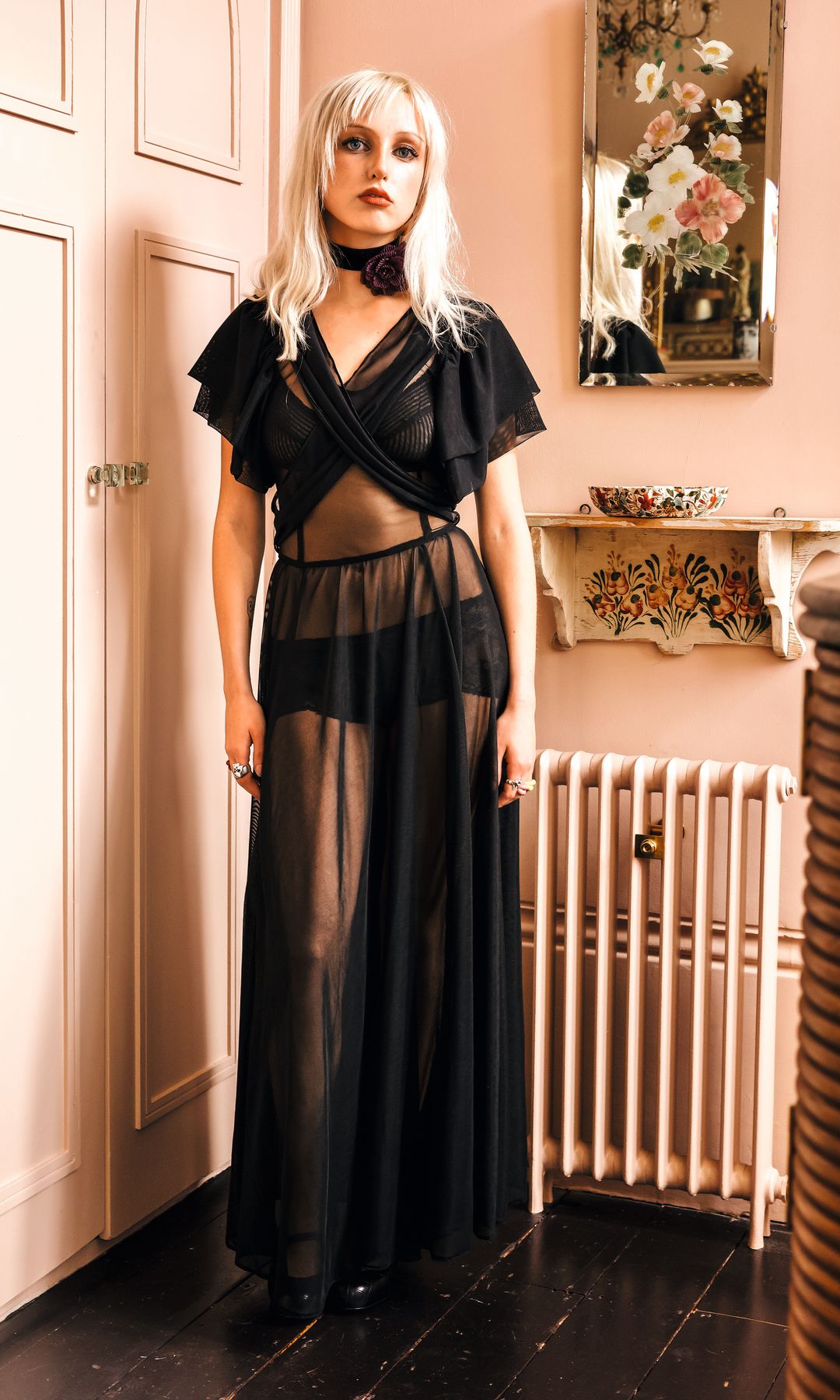 Mujer con cabello rubio largo, vestida con un vestido negro transparente, posando en una habitación con un radiador y un espejo decorativo.