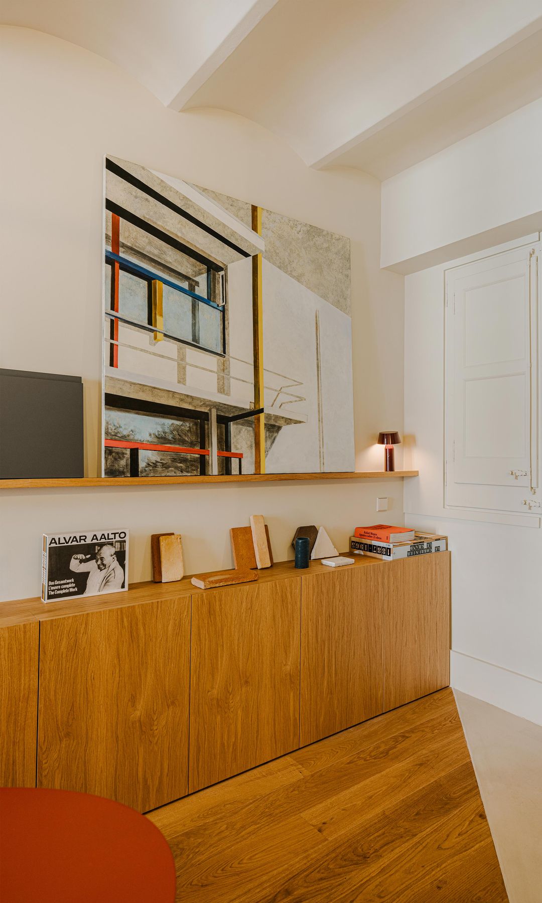 Rincón comedor con mesa roja, suelo de madera, cortinas, aparador de madera con puertas y libros en la repisa, esntería de madera con lámpara de sobremesa roja