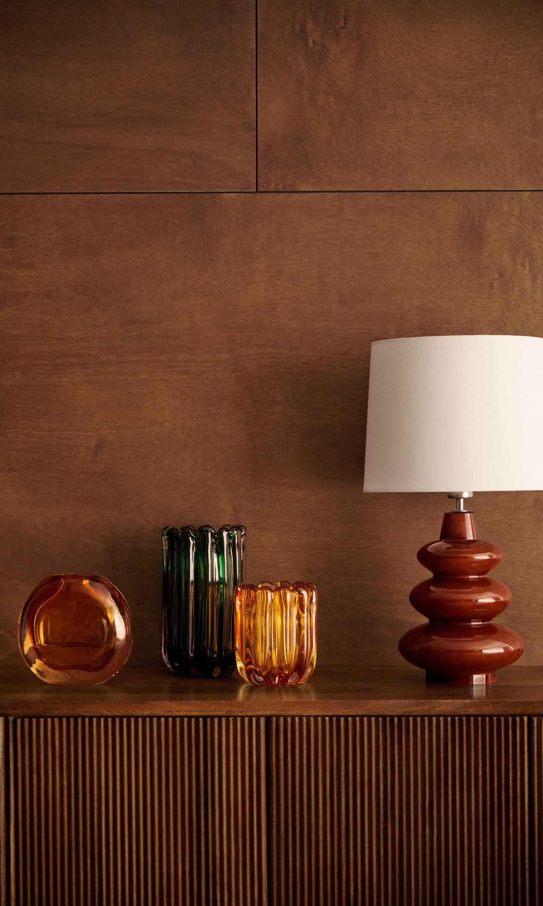 Pared de madera oscura y aparador con lámpara de sobremesa con el pie marrón y la pantalla blanca y jarrones en color ámbar