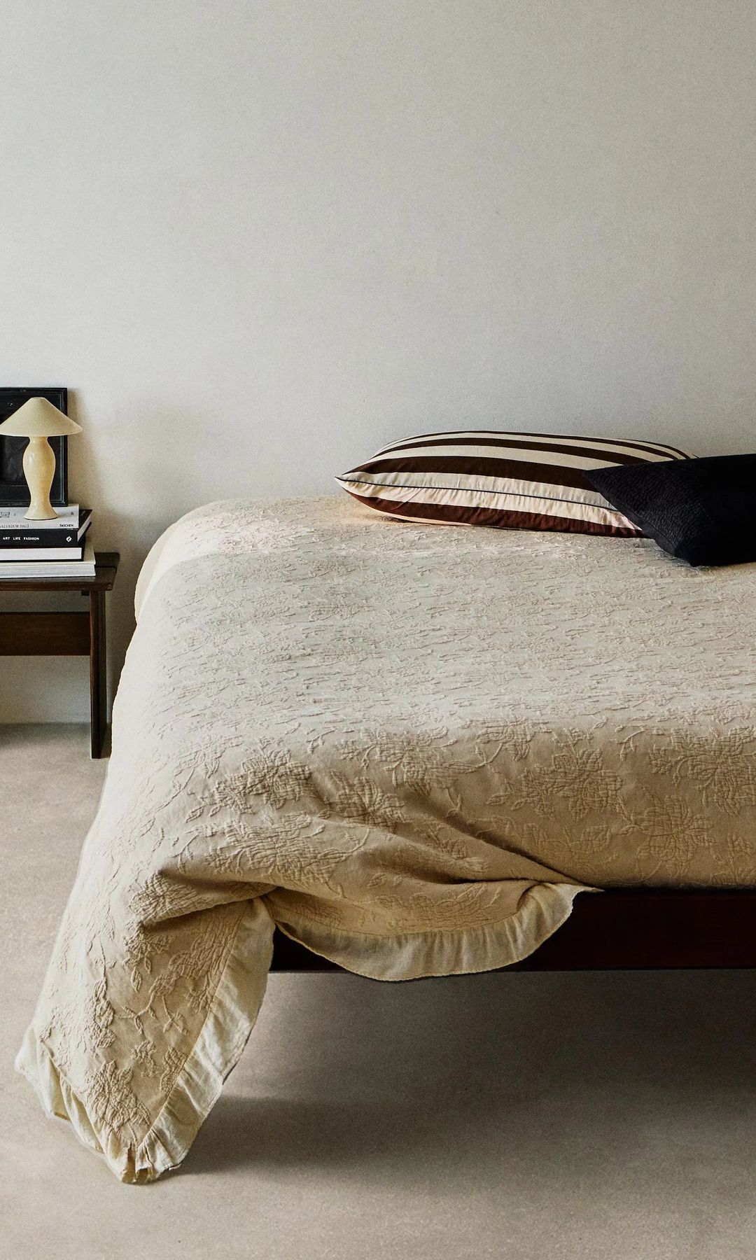 Colcha bordada de Zara Home con cojines negros, mesita de noche de madera oscura, lámpara de sobremesa 