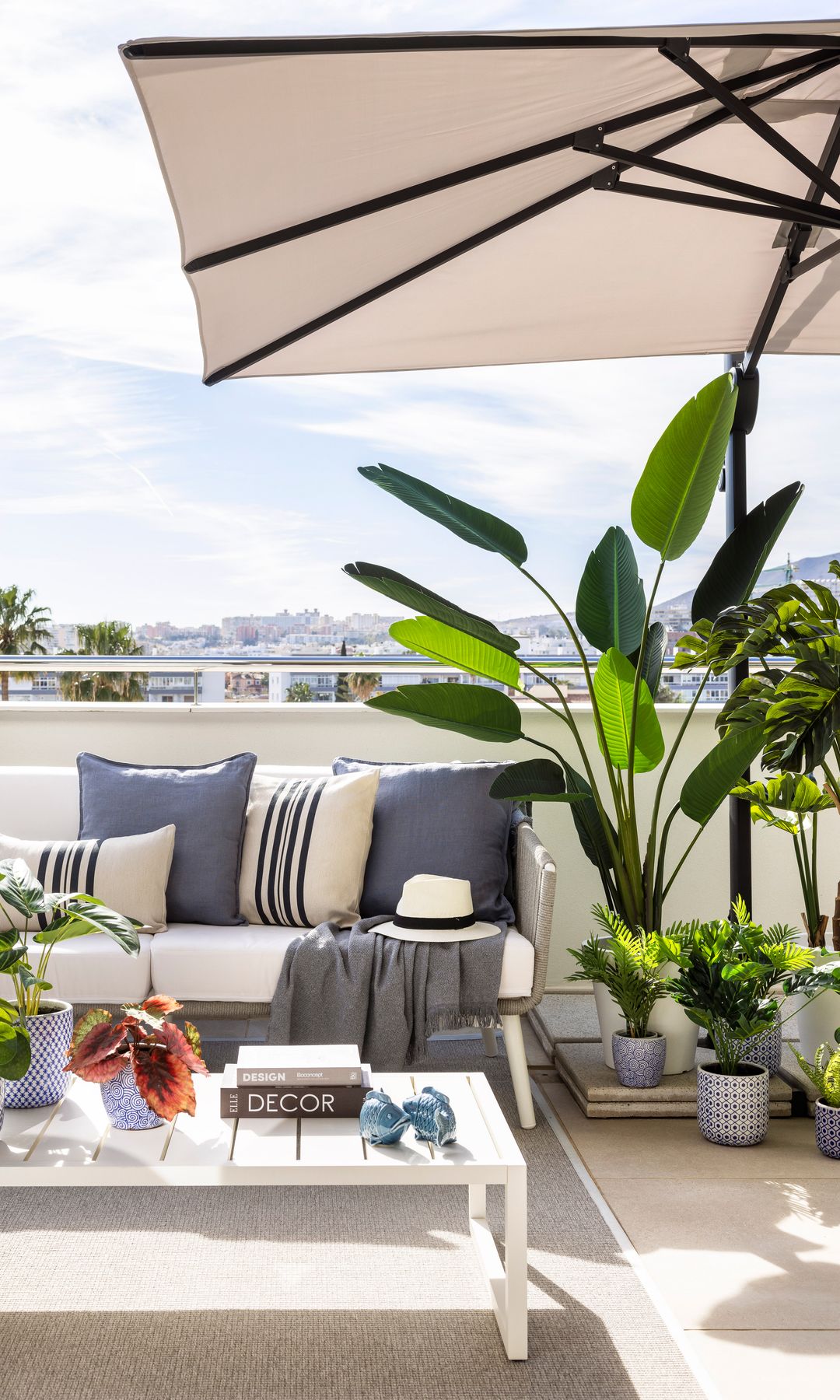 Terraza con sombrilla blanca, sofá blanco, cojines azules y blancos de rayas, macetas con plantas altas
