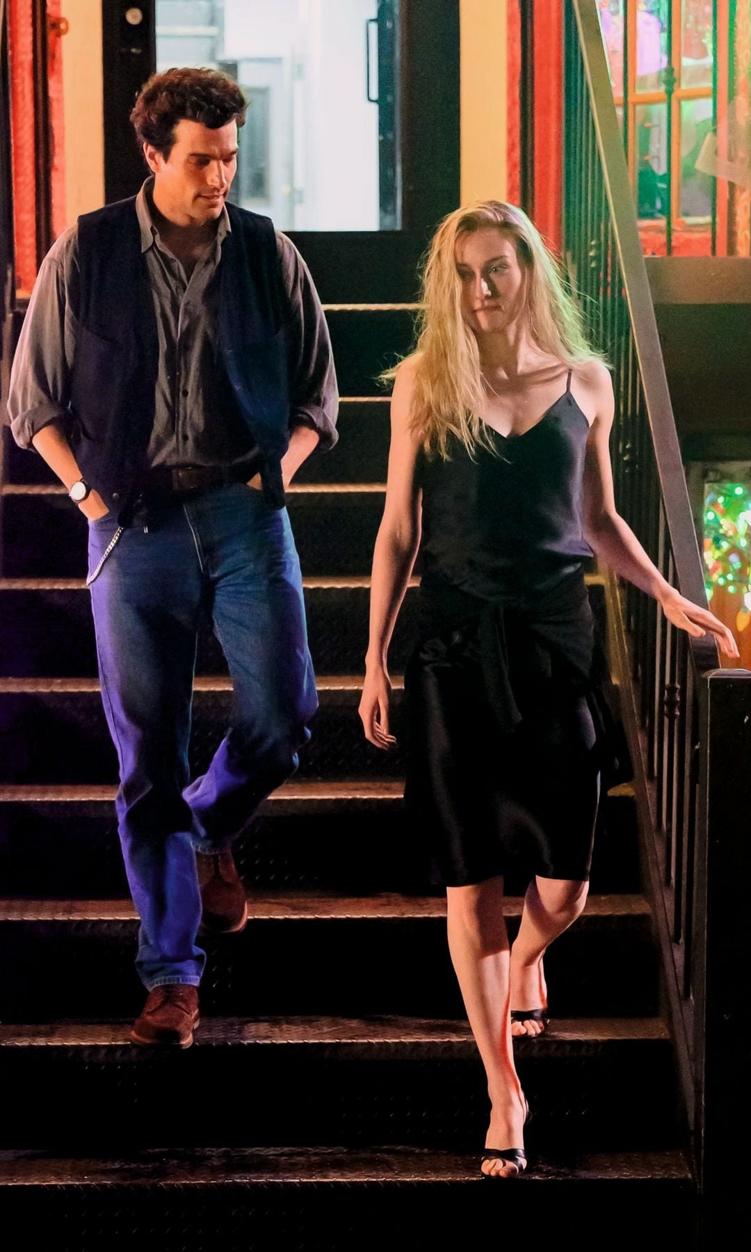 Hombre y mujer bajando escaleras. Él en jeans y chaleco, ella en vestido negro.