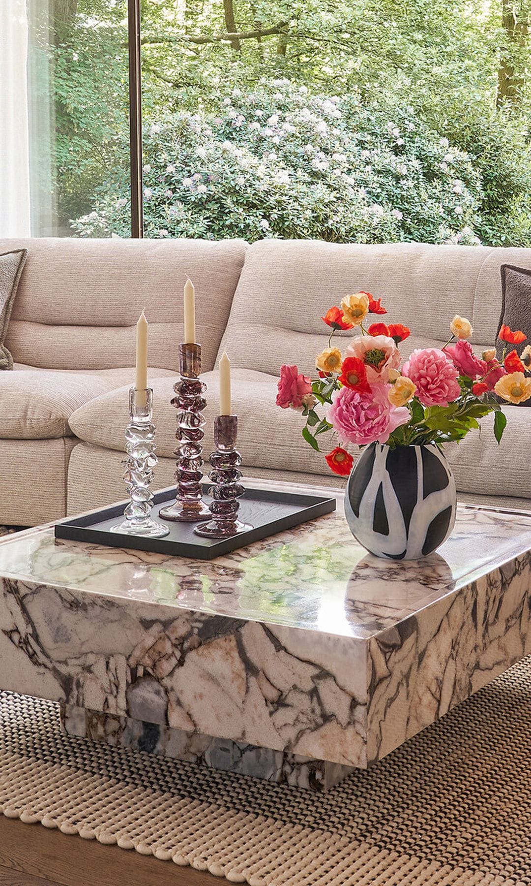 Salón con sofá beige, mesa de centro de mármol con jarrón blanco y negro con flores de colores y bandeja con candelabros 