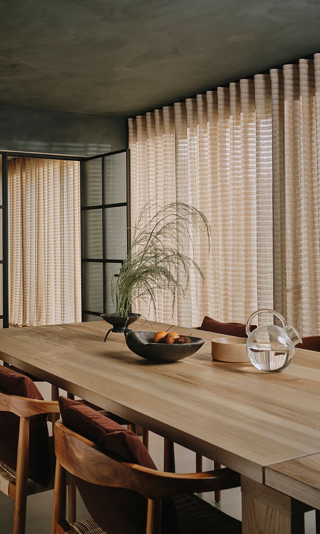 Comedor con mesa XL de madera, sillas de madera, jarrón y frutero sobre la mesa y cortinas beiges colgadas desde el techo 