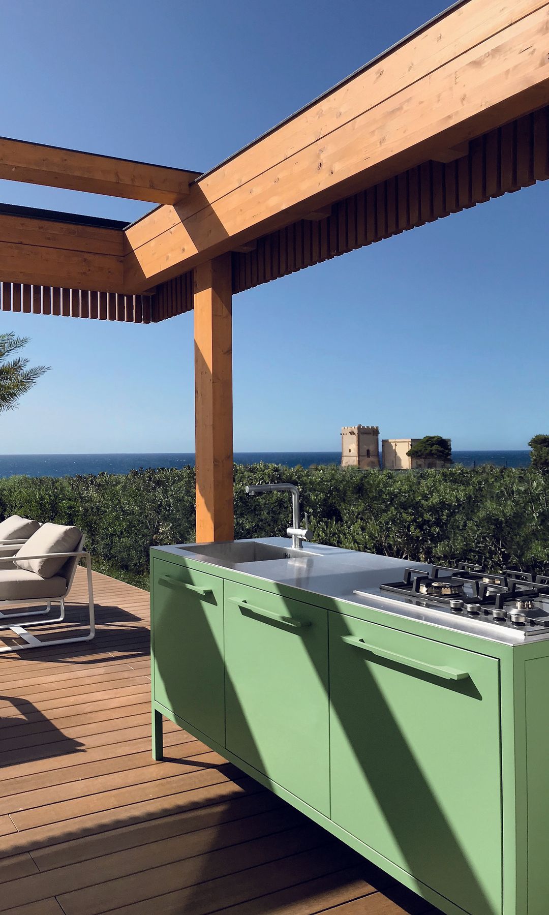Cocina exterior verde con fuegos y fregadero bajo pérgola de madera, suelo de madera