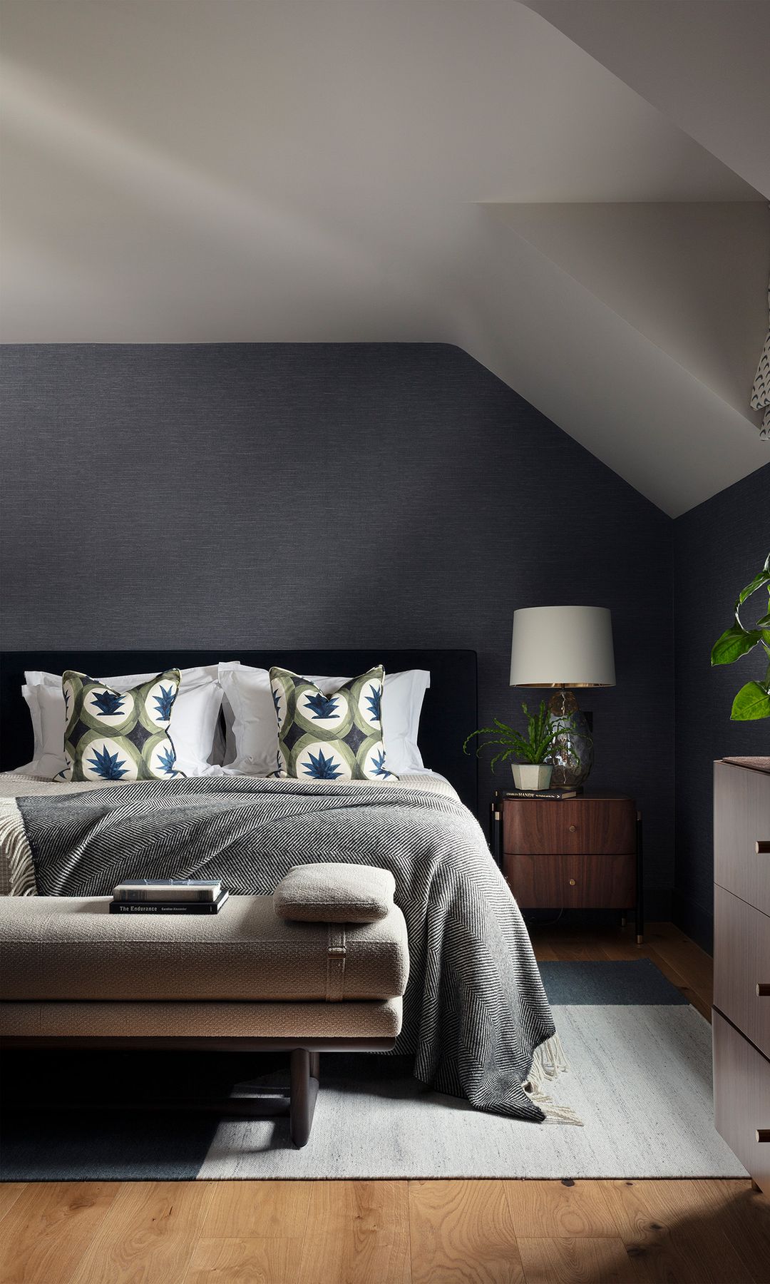 Dormitorio abuhardillado con paredes grises, ropa de cama gris con cojines verdes, cómoda de madera, plantas, lámpara de sobremesa, mesita de noche, banqueta tapizada