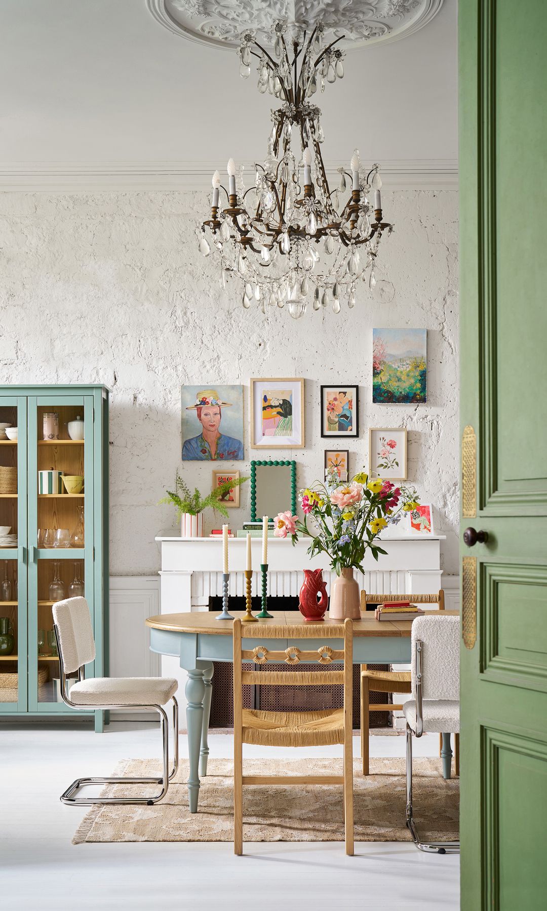 Comedor con chimenea, puerta verde, mesa de comedor vintage con las patas torneadas de color azul claro y el sobre de madera, sillas de comedor diferentes, lámpara de techo de cristal, cuadros en las paredes, vitrina de color azul claro, jarrón con flores rosas